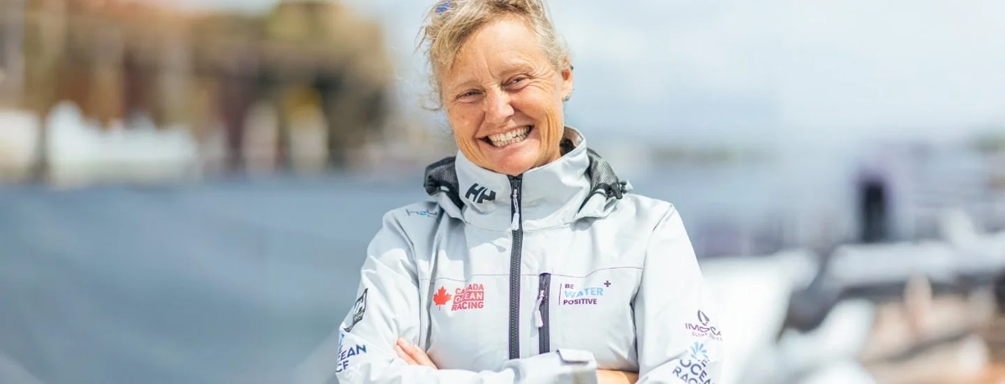 Smiling woman in a white jacket outdoors with boats in the background.