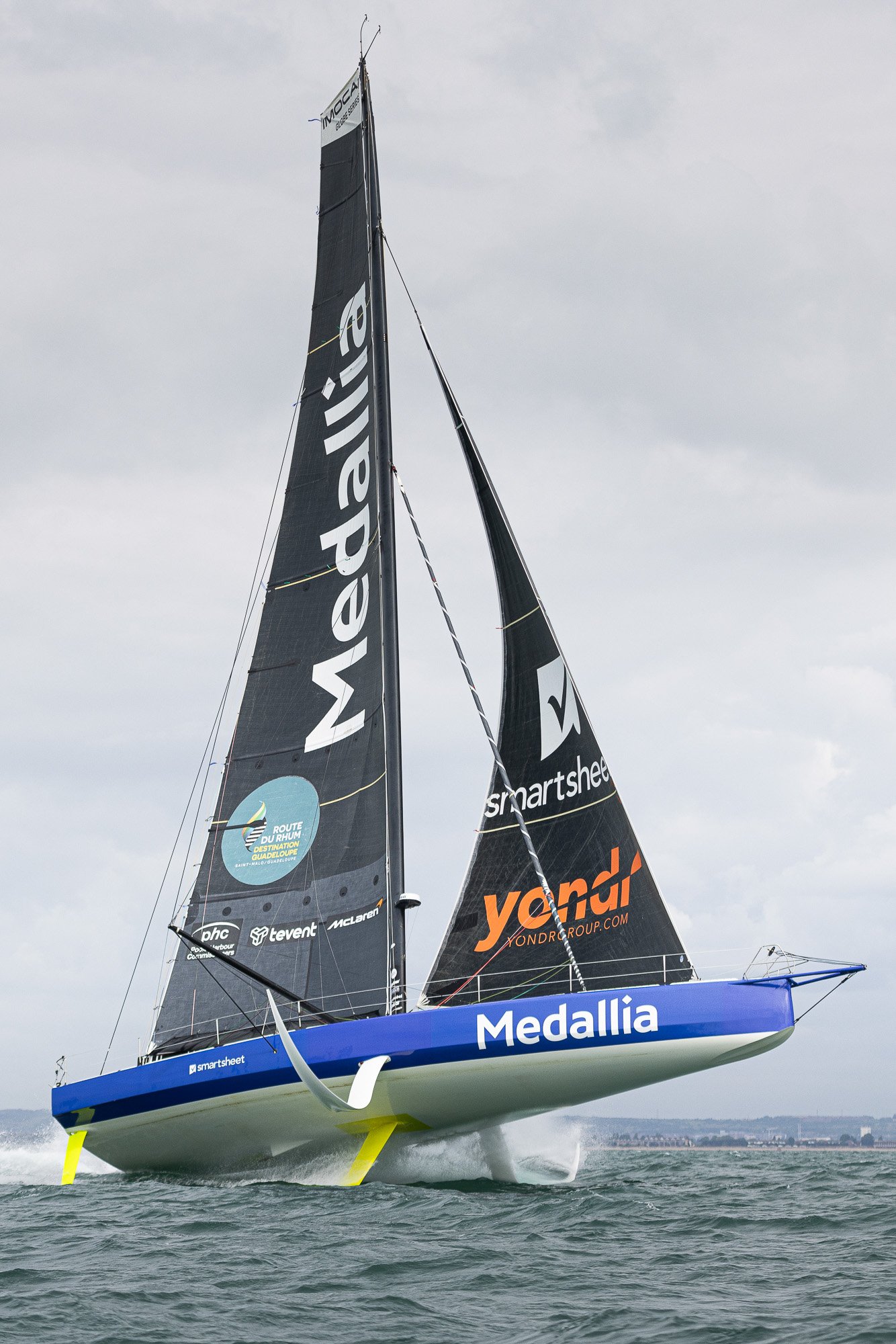 Pip Hare Ocean Racing offshore racing yacht with black sails and Medallia branding sailing at speed under a cloudy sky, with foils lifting the hull clear of the water.