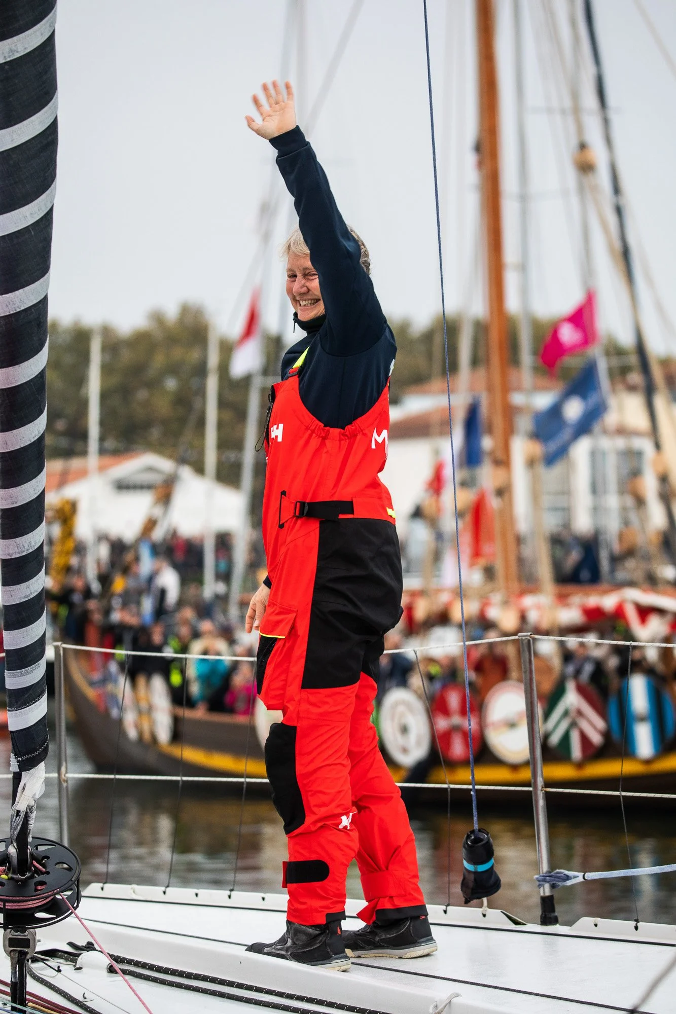 Pip Hare Ocean Racing sailor waving from bow of sailboat, with a crowd and moored boats in the background. Vendee Globe.