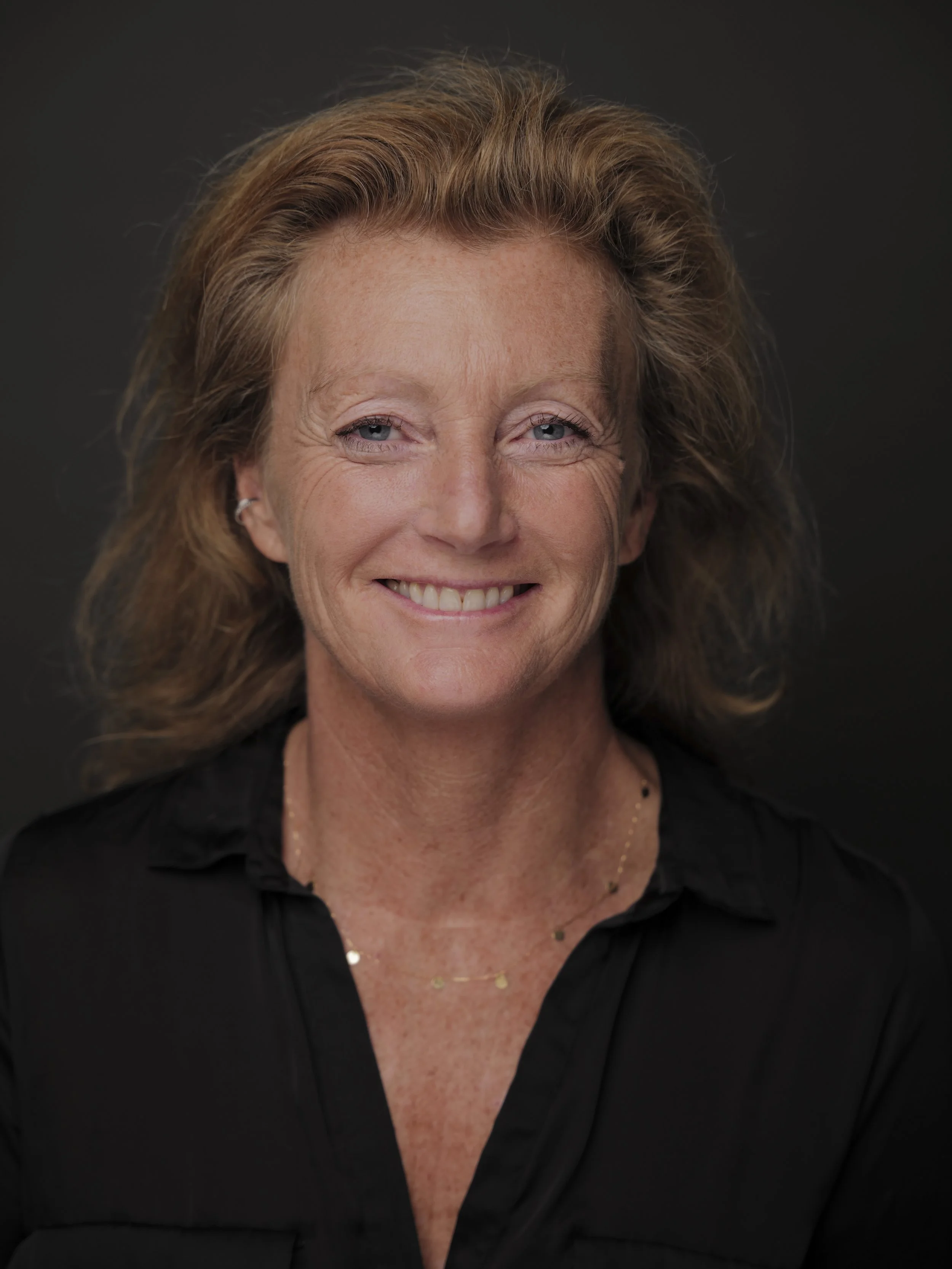 Photo d'une femme souriante avec des cheveux roux, portant une chemise noire et un collier fin, contre un fond sombre.