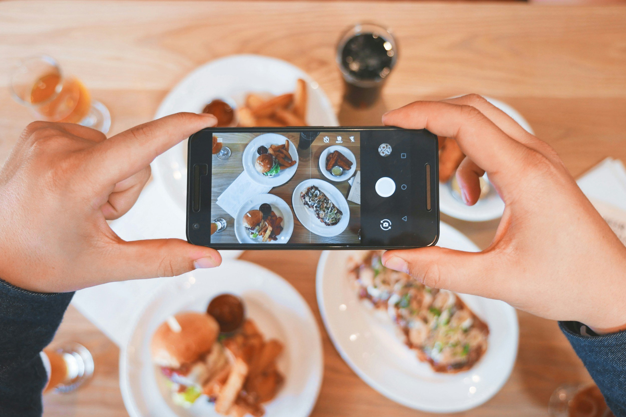 Une personne prend une photo de plats de nourriture variée à l'aide d'un smartphone. La table contient plusieurs assiettes avec différents plats, notamment des burgers, des sushi et des frites. Il y a aussi des verres et des boissons.