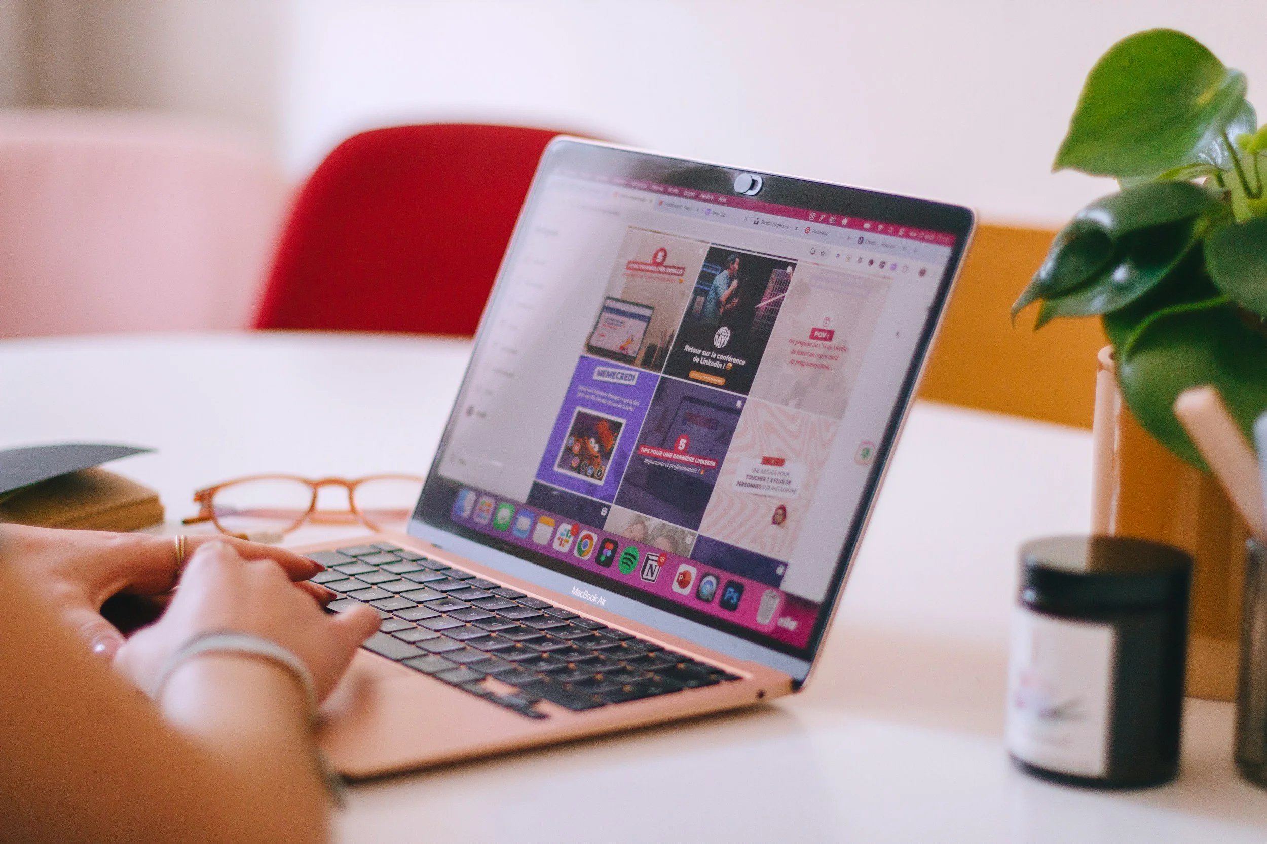 Une personne utilisant un ordinateur portable rose sur un bureau, avec des lunettes orange, un carnet et une plante verte à proximité.