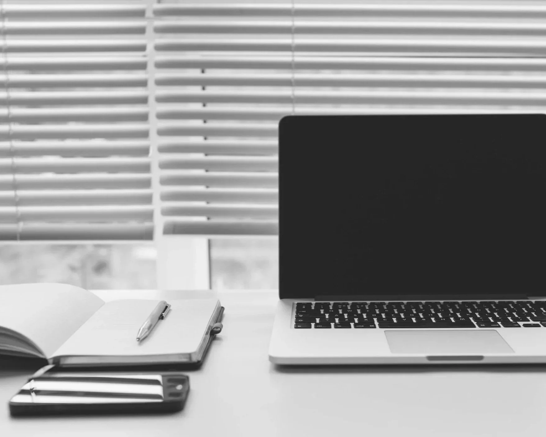 A workspace featuring a laptop, an open notebook with a pen, a closed smartphone, and a window with blinds in the background.