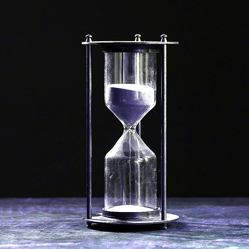 An hourglass with clear glass and black sand, standing on a dark wooden surface against a black background.