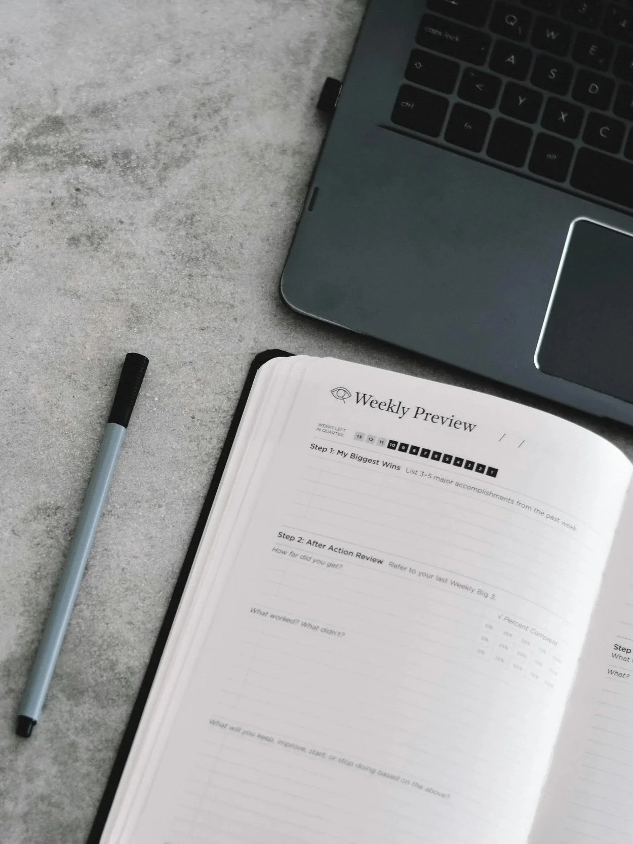 Open notebook titled Weekly Preview on gray surface with black pen and gray laptop