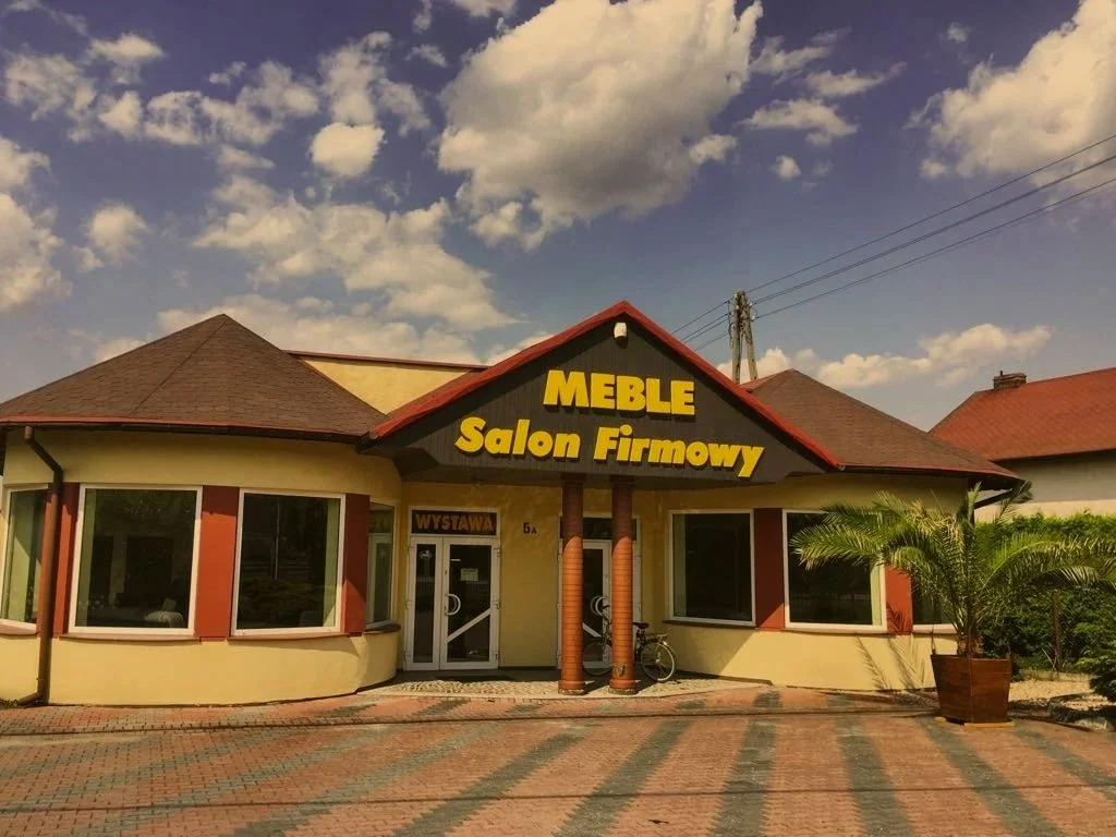 A yellow building with a brown roof, featuring a large sign that reads "MEBLE Salon Firmowy" in yellow letters on a black background. There are bay windows on either side of a glass door, with a bicycle parked outside and a potted palm tree to the right.