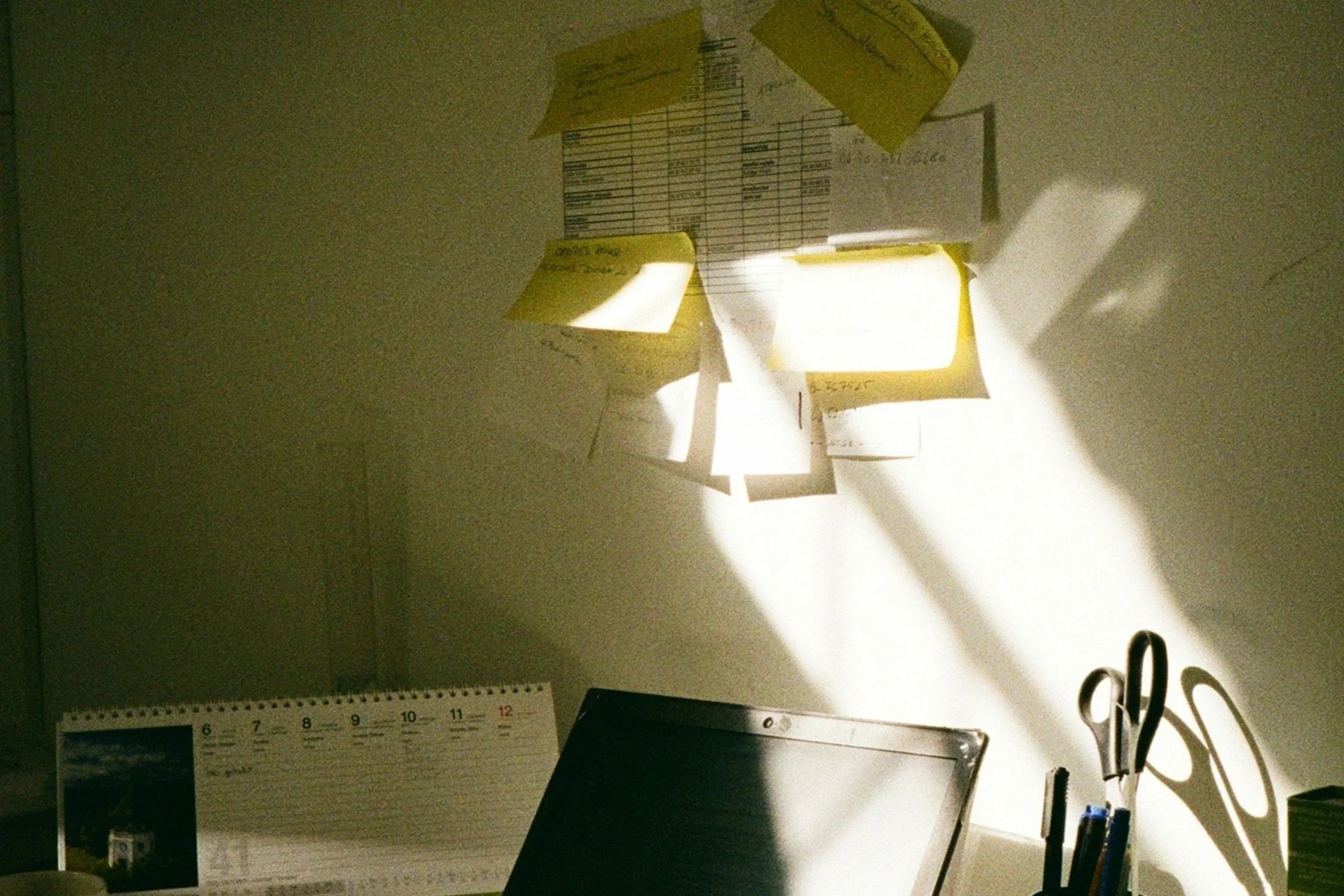 Espace de bureau avec un calendrier sur une table, une paire de ciseaux et des stylos dans un porte-stylo, et des notes adhésives collées au mur. La lumière crée des ombres sur le mur.