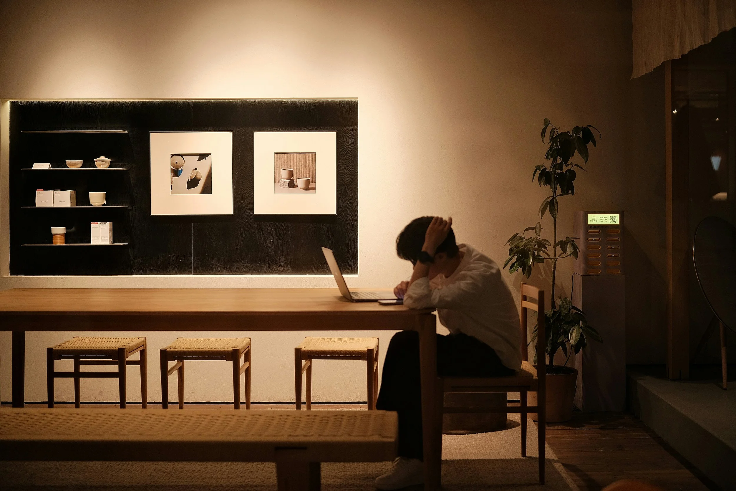 Une personne assise à une table en bois, la tête dans les mains, devant un ordinateur portable dans une pièce tamisée. ©ntérieur de style minimaliste avec une étagère noire, des œuvres d'art, une plante verte et un dispositif électronique à l’arrière.