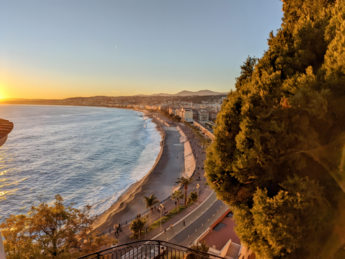Vue d'une côte avec plage, mer, route, palmiers, arbres et buildings au coucher du soleil.