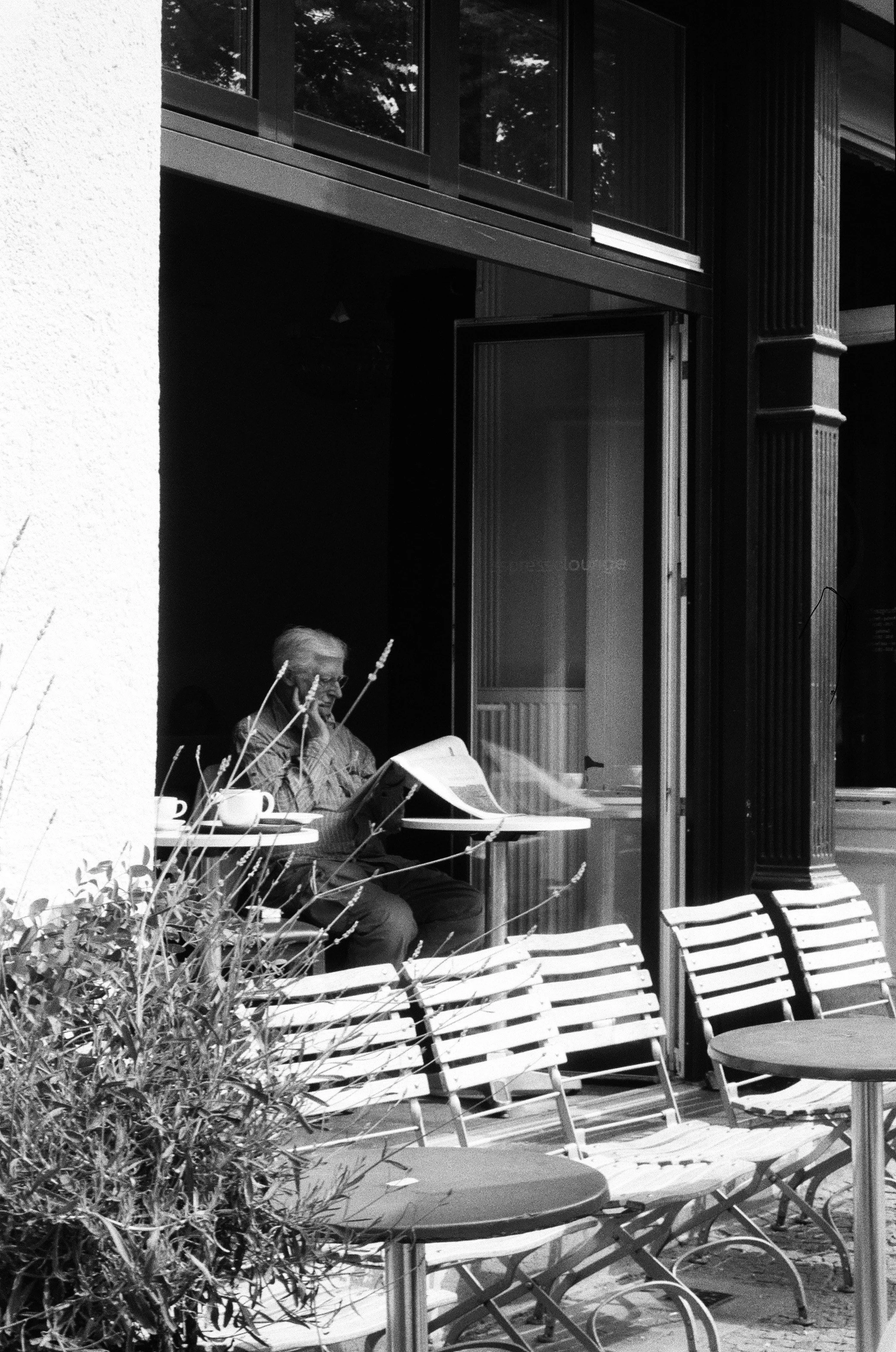 Un homme âgé lisant un journal à une table en terrasse devant un bâtiment avec de grandes fenêtres en verre.
