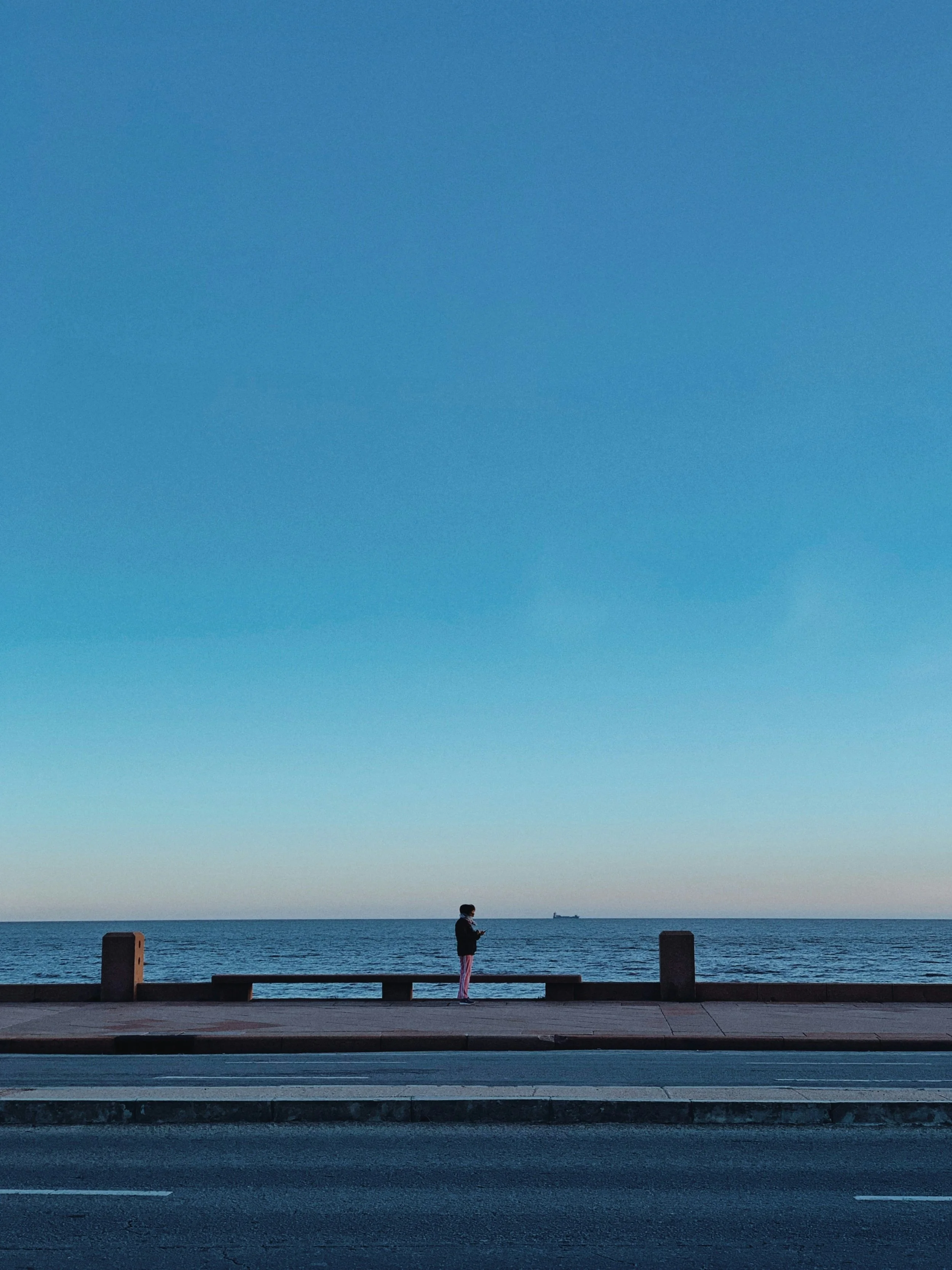 Une personne se tient sur un banc en regardant la mer, avec un bateau en arrière-plan, sous un ciel clair.