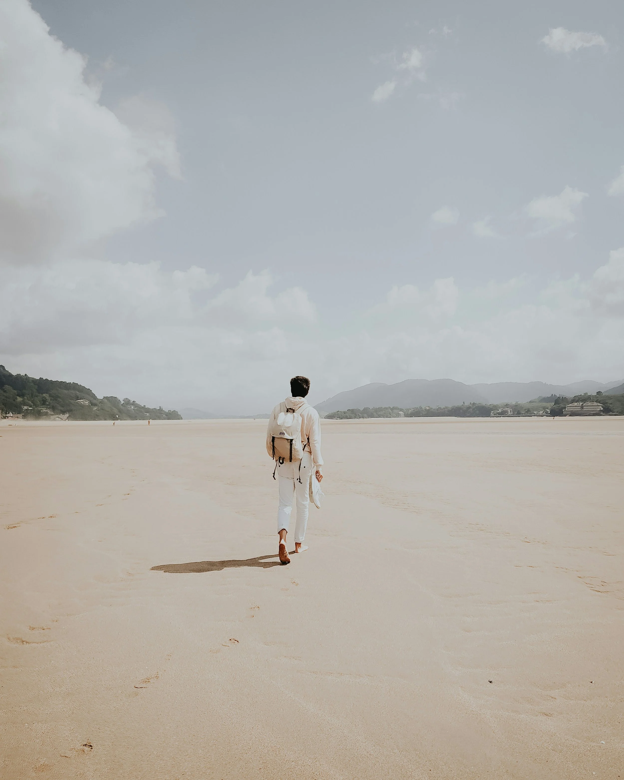Un homme marche seul sur une plage de sable avec une montagne en arrière-plan, ciel nuageux.