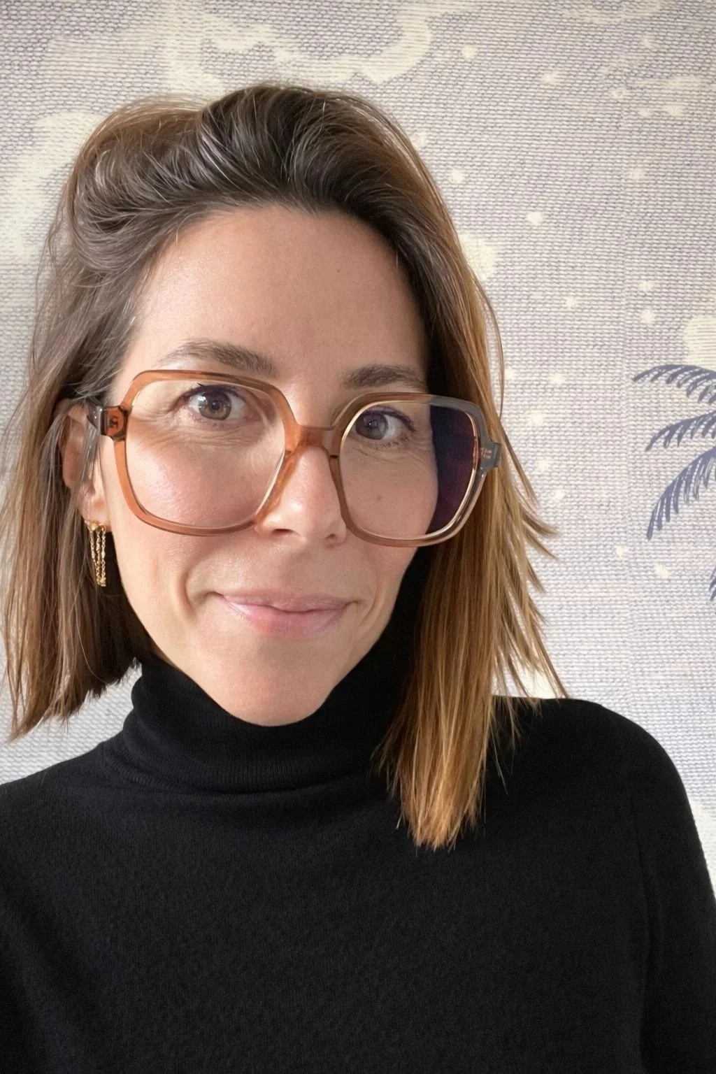 Une femme avec des cheveux bruns courts, portant des lunettes épaisses orange, un haut noir à col roulé, et des boucles d'oreilles dorées, devant un fond clair avec des motifs de feuilles et de points.