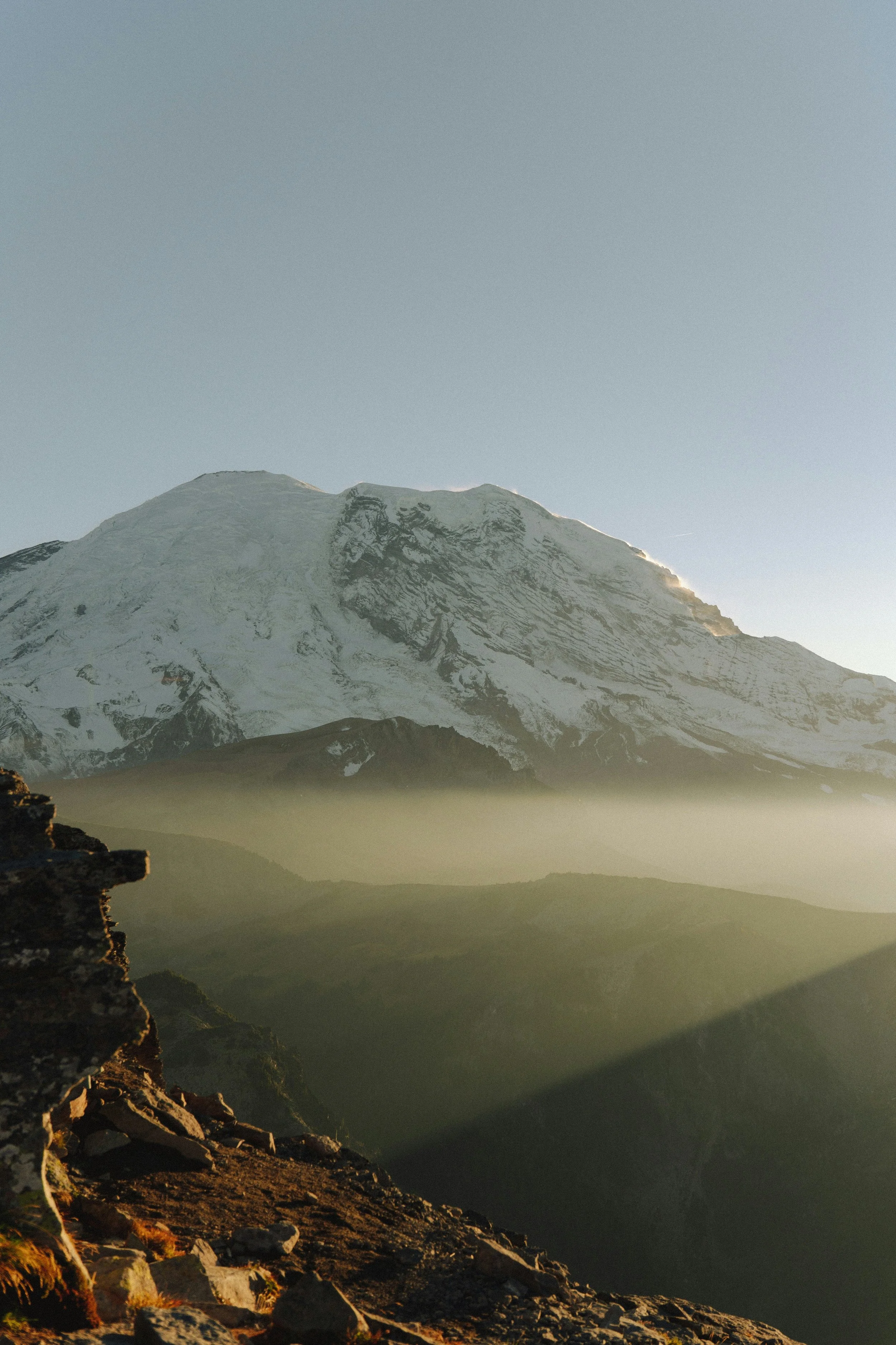 Montagne enneigée vue au lever ou coucher du soleil, avec un premier plan rocheux et des couches de brume ou de nuages basses.