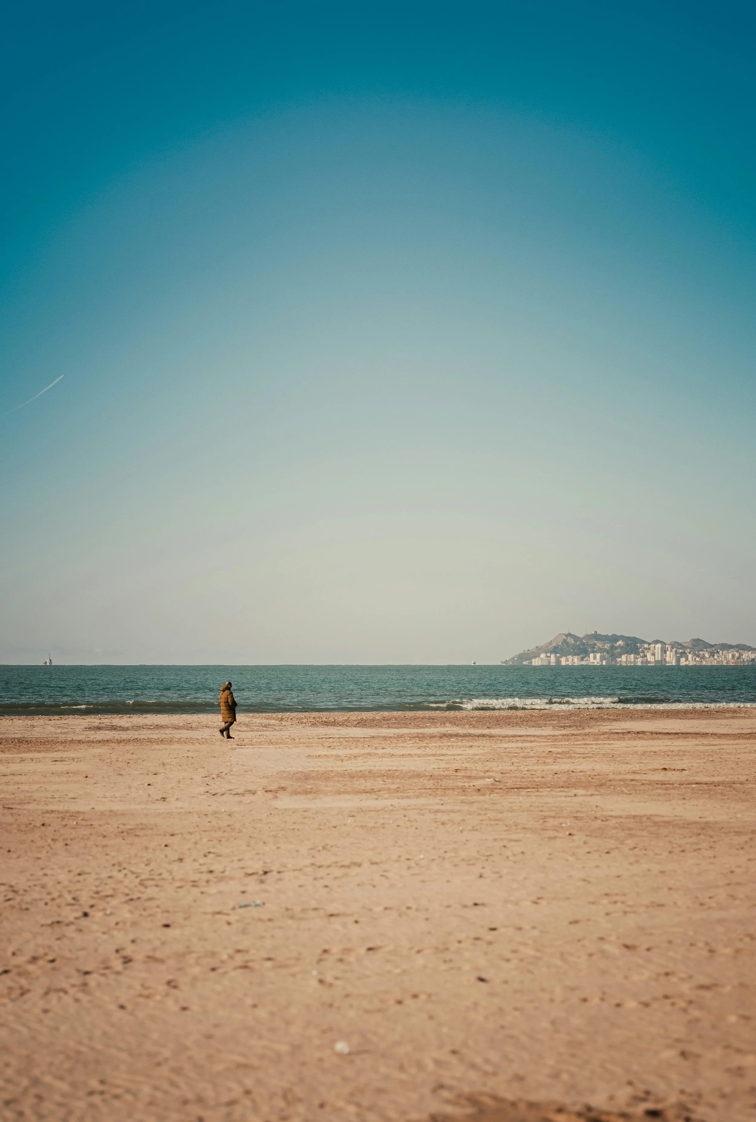 Une personne marche seule sur une plage de sable, avec la mer à l'arrière-plan et une ville visible au loin sur la droite.
