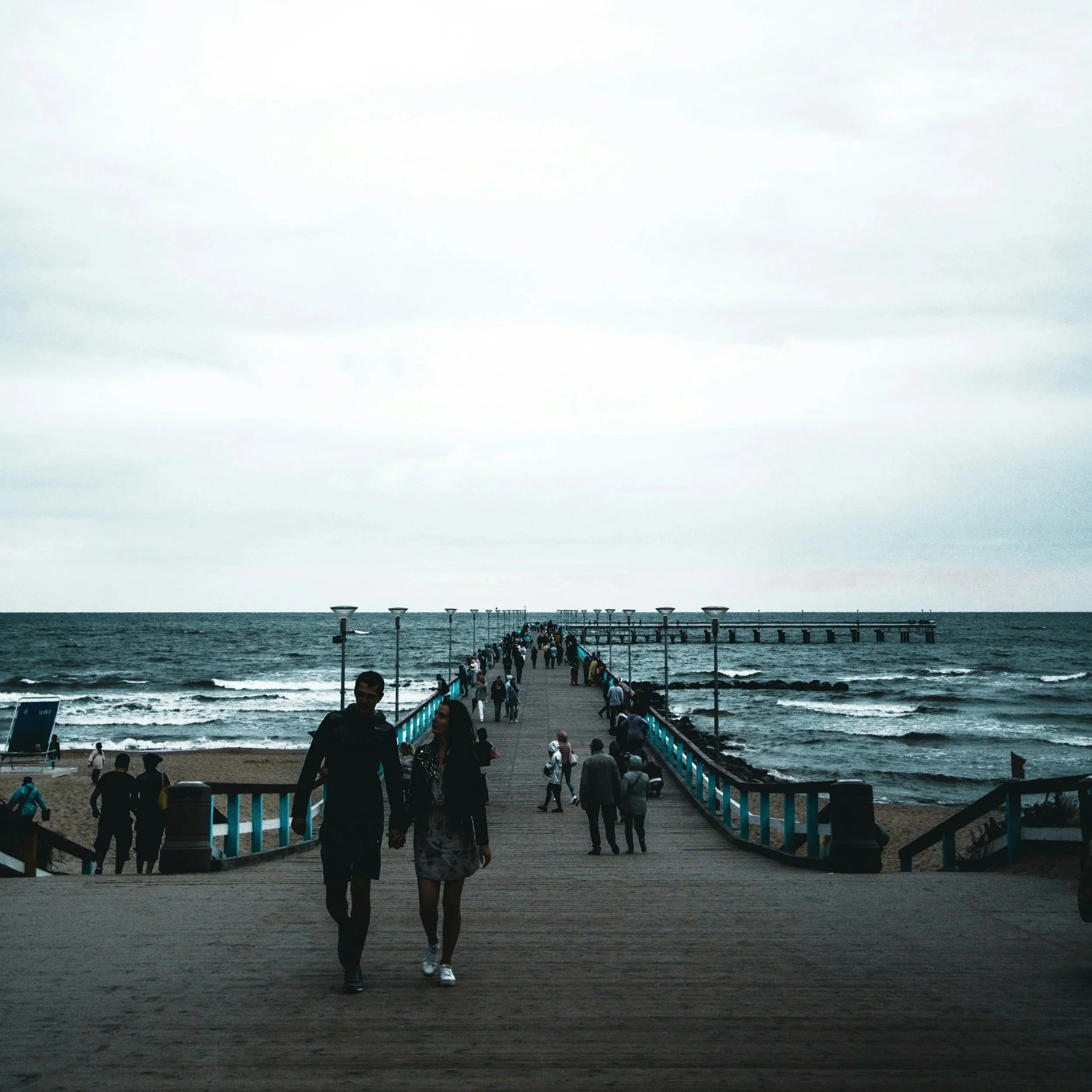 Une jetée en bois s'étendant dans la mer avec des personnes marchant dessus, sous un ciel nuageux.