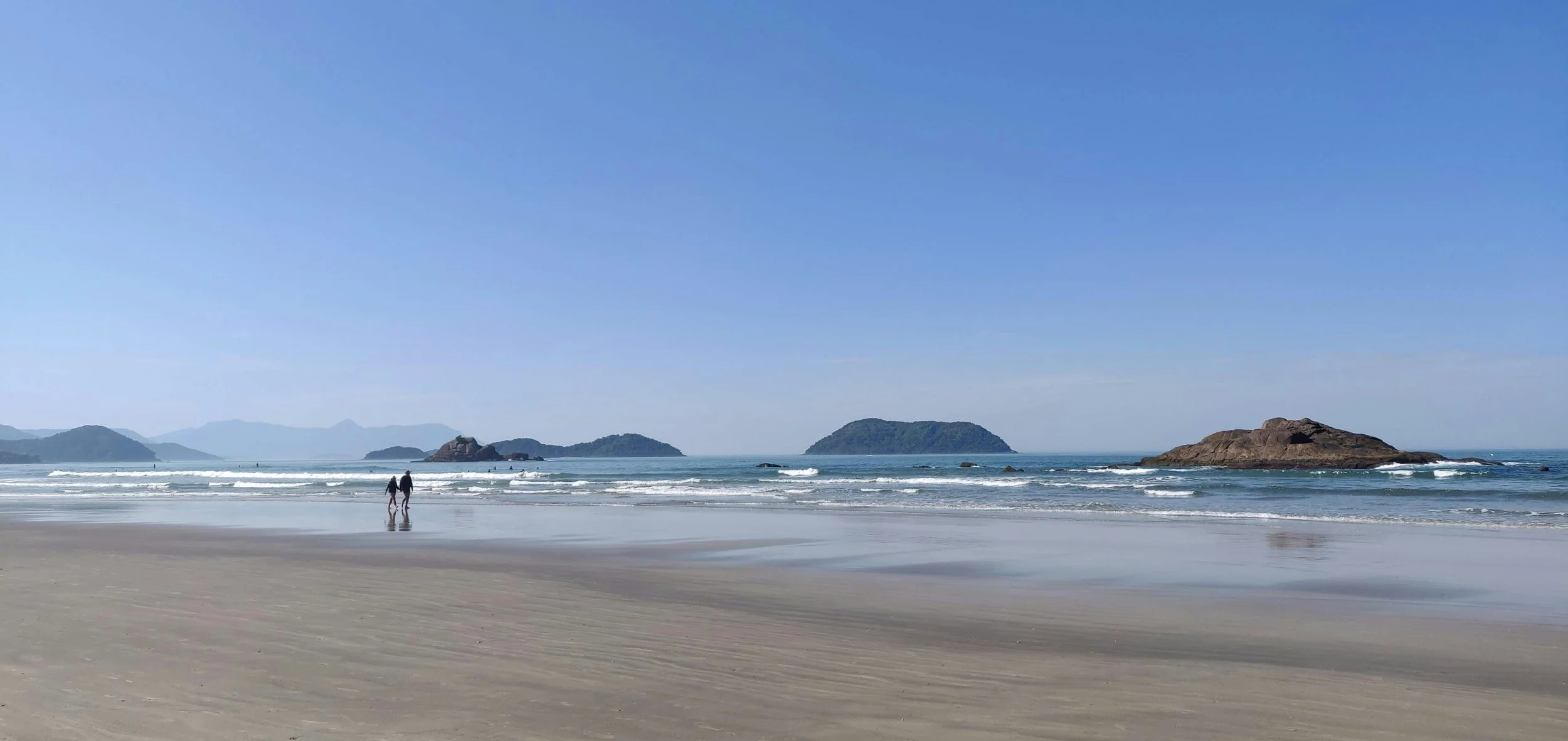 Plage de sable avec deux personnes marchant près de l'eau, plusieurs îles rocheuses au large, ciel clair et horizon flou dans un paysage marin.