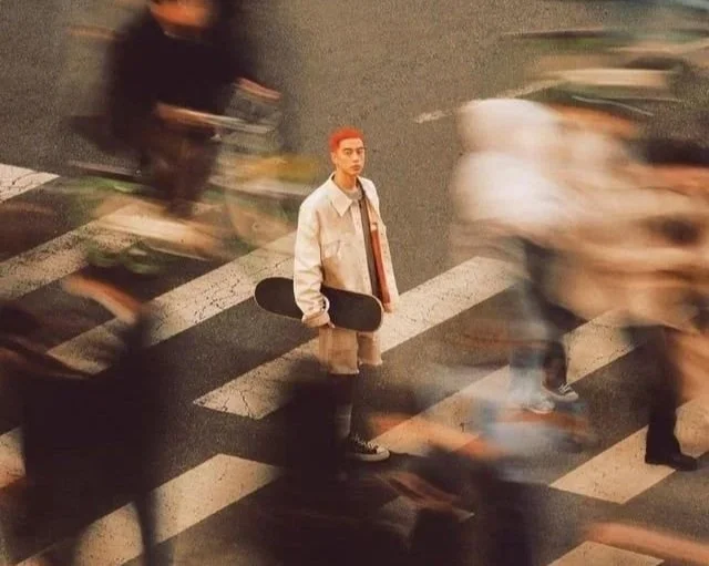 Jeune homme avec un skateboard, debout au centre d'un passage piéton, entouré de personnes en mouvement.