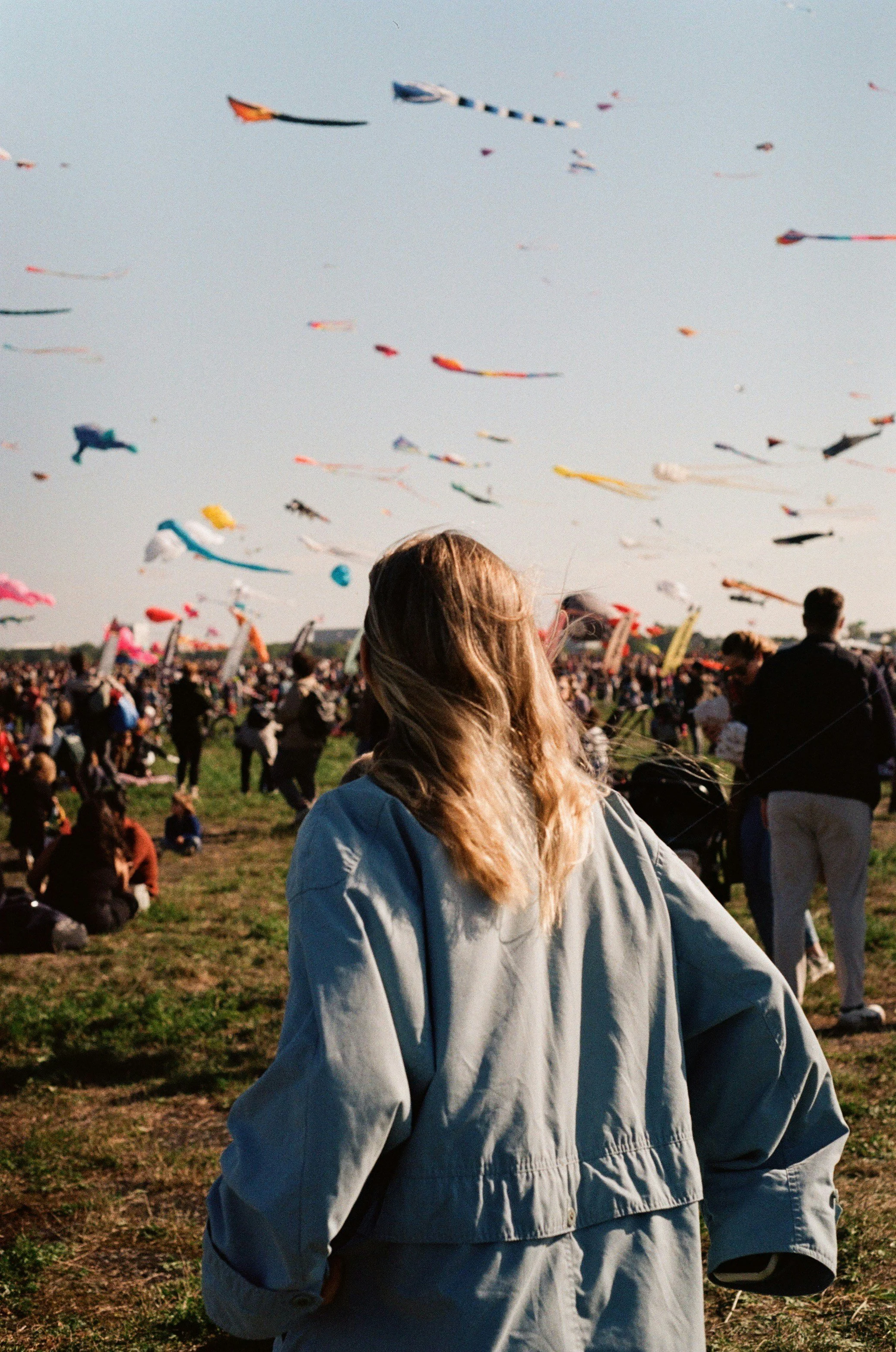 Une femme vue de derrière assiste à un festival en plein air avec des cerfs-volants colorés dans le ciel, beaucoup de personnes présentes autour dans un grand champ.