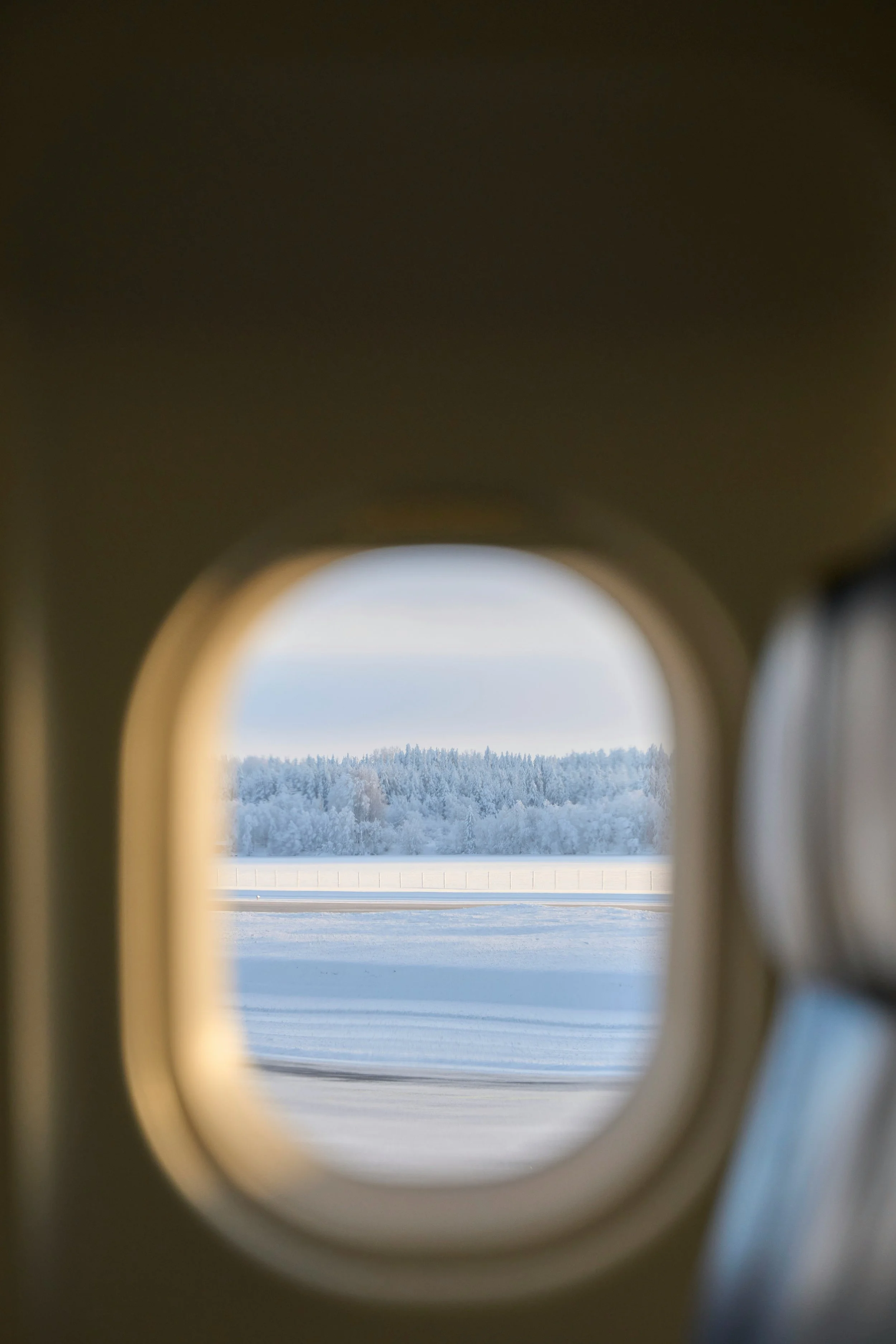 Paysage hivernal vu à travers une petite fenêtre d'avion, avec des arbres recouverts de neige et un ciel clair.