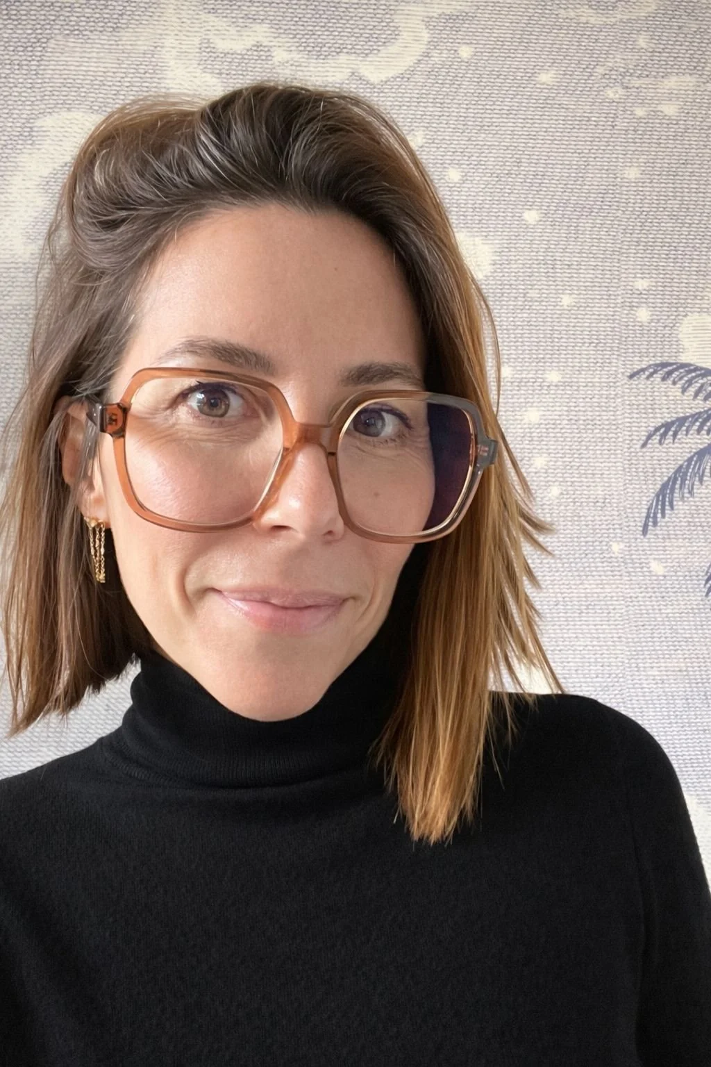 Une femme portant des lunettes épaisses teintées de couleur orange, un haut noir à col roulé, avec des boucles d'oreilles dorées, devant un fond clair avec un dessin de palmier.