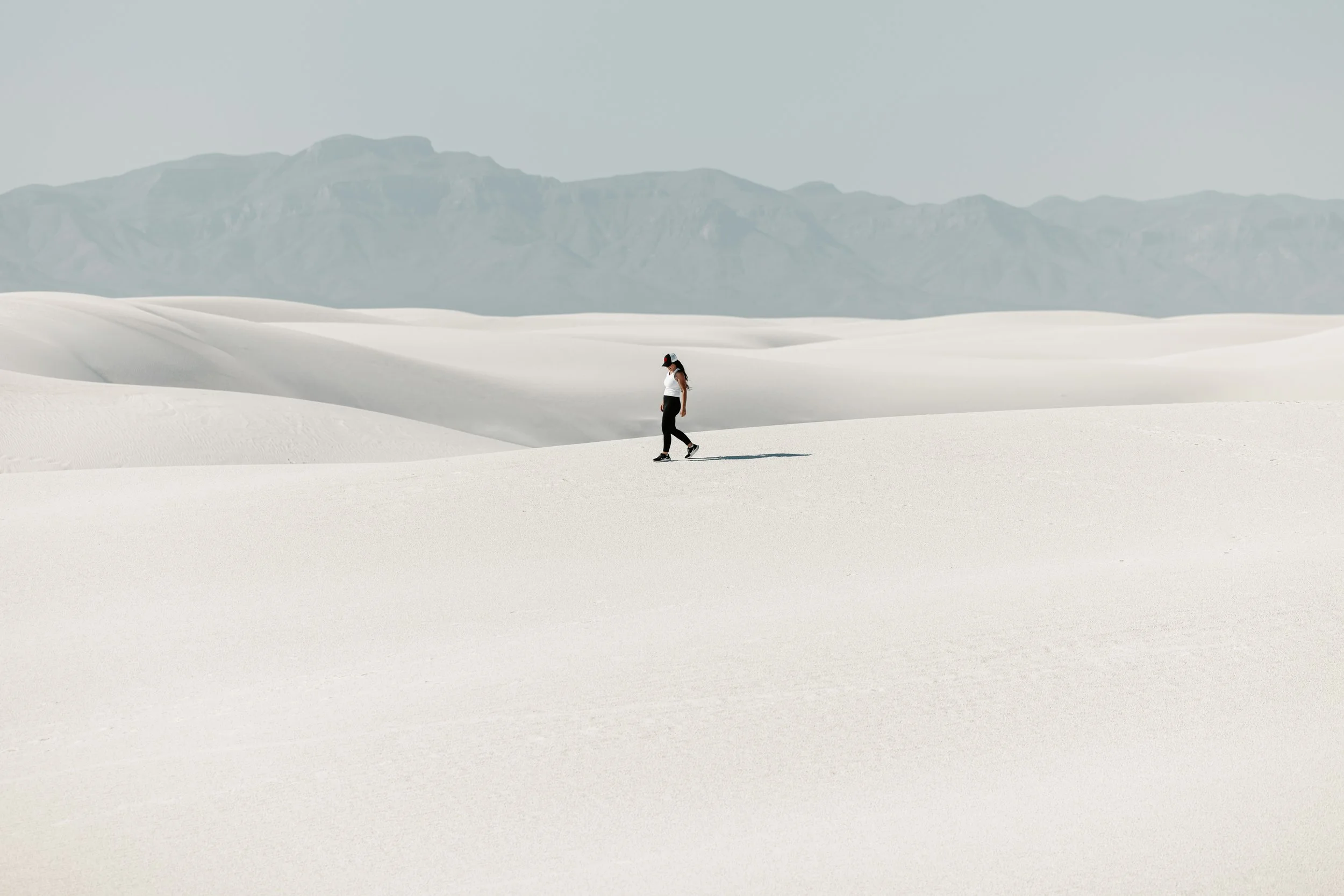 Une femme portant un chapeau, une t-shirt blanc et un pantalon noir marche dans un désert de dunes de sable blanc, avec des montagnes en arrière-plan.