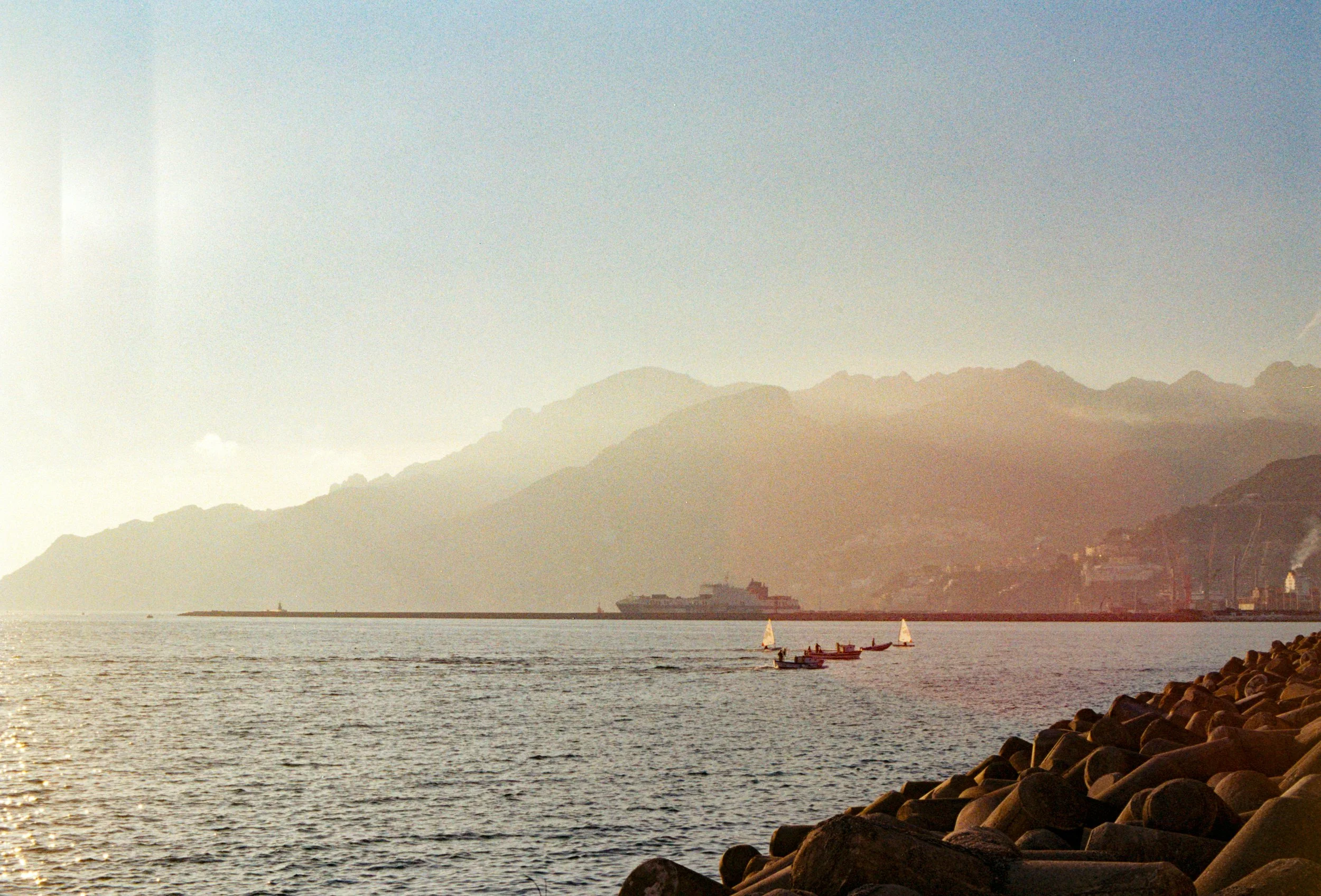 Scène maritime avec des bateaux de pêche en eaux calmes, montagne en arrière-plan, ciel clair et roche au premier plan.