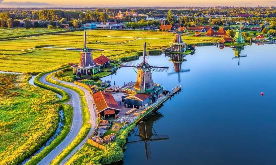 Aerial view of traditional Dutch windmills along a river with green fields and farm buildings in the background at sunset.