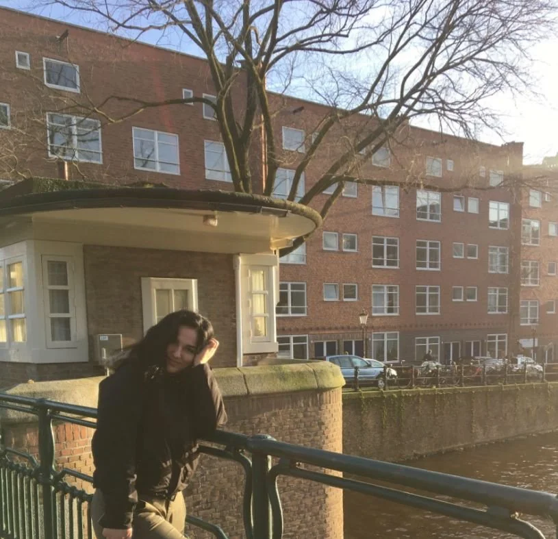 A woman leaning on a railing next to a body of water with an apartment building in the background during sunset.