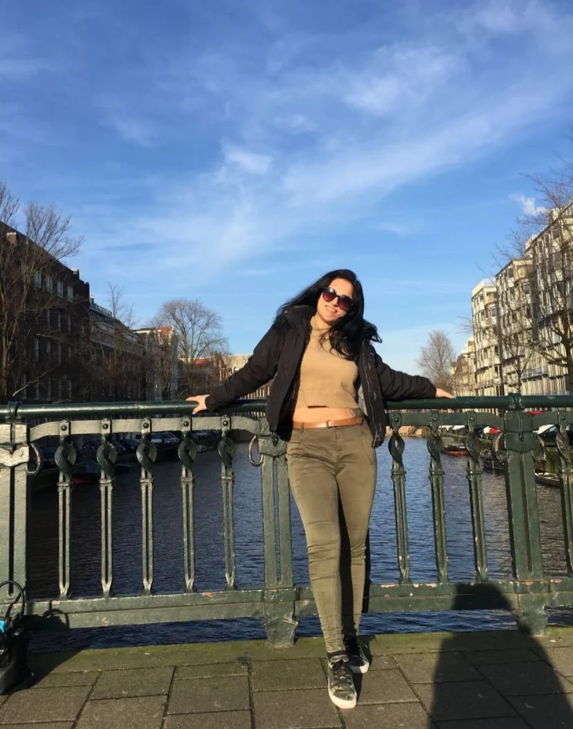 Woman posing on a footbridge over a canal with apartment buildings and leafless trees in the background under a partly cloudy blue sky.