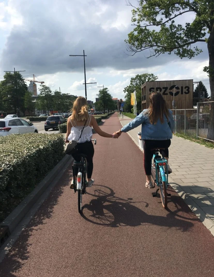 Two women riding bicycles hand-in-hand on a city bike path on a cloudy day, with parked cars on the side and trees lining the path.