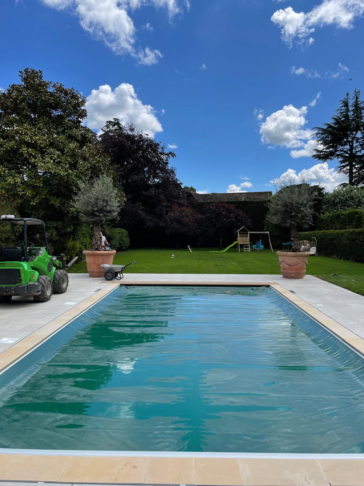 Residential backyard with a rectangular swimming pool covered with a safety cover, surrounded by a light-colored tile patio. In the background, there are lush green grass, large potted trees, garden toys, a small slide, a swing set, and outdoor seating. The sky is partly cloudy with scattered white clouds.