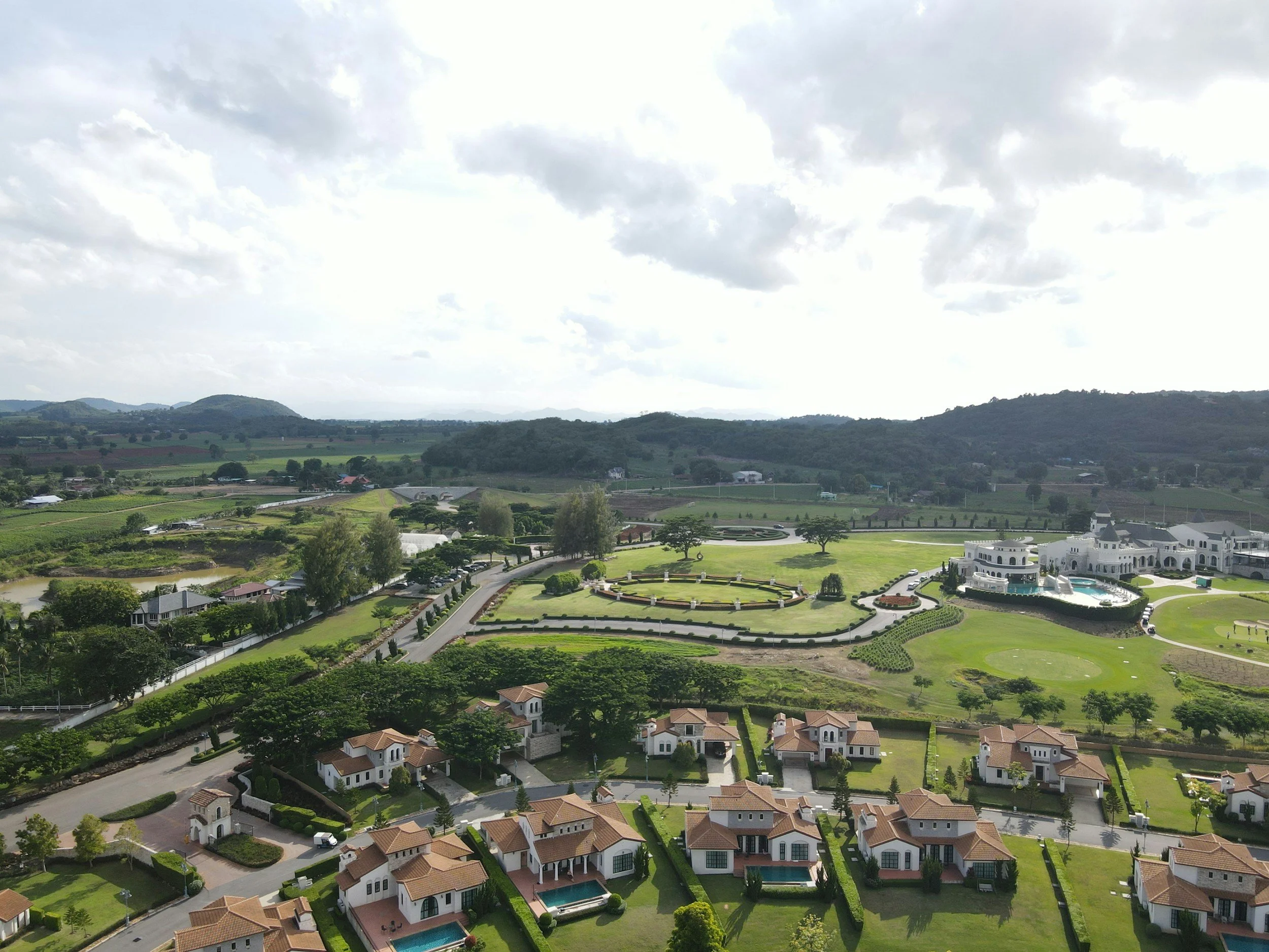 Aerial view of a luxury residential community with white houses, green lawns, and a large estate with a mansion and pool, surrounded by hills and farmland under a cloudy sky.