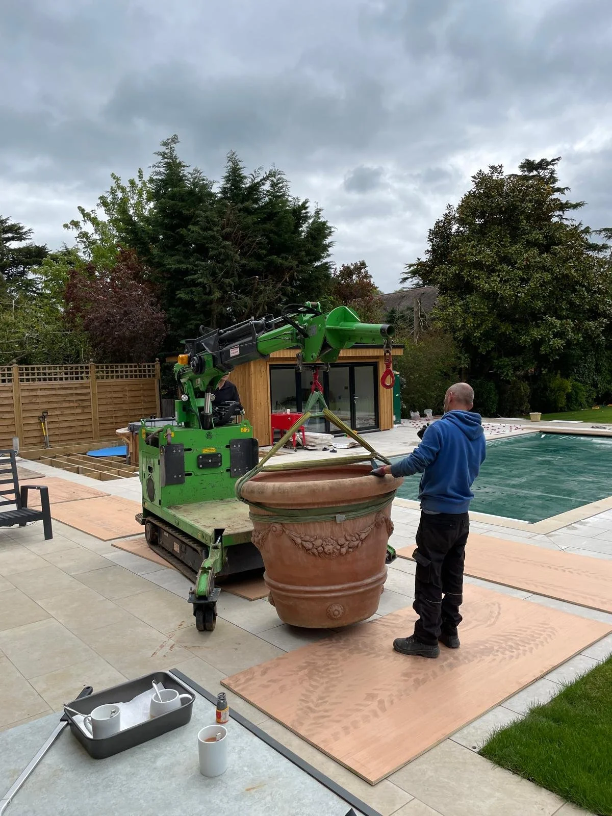 A man operating a green crane lifts a large terracotta planter with decorative carvings in a backyard. There is a swimming pool, a wooden shed, and a wooden fence in the background. The sky is cloudy.