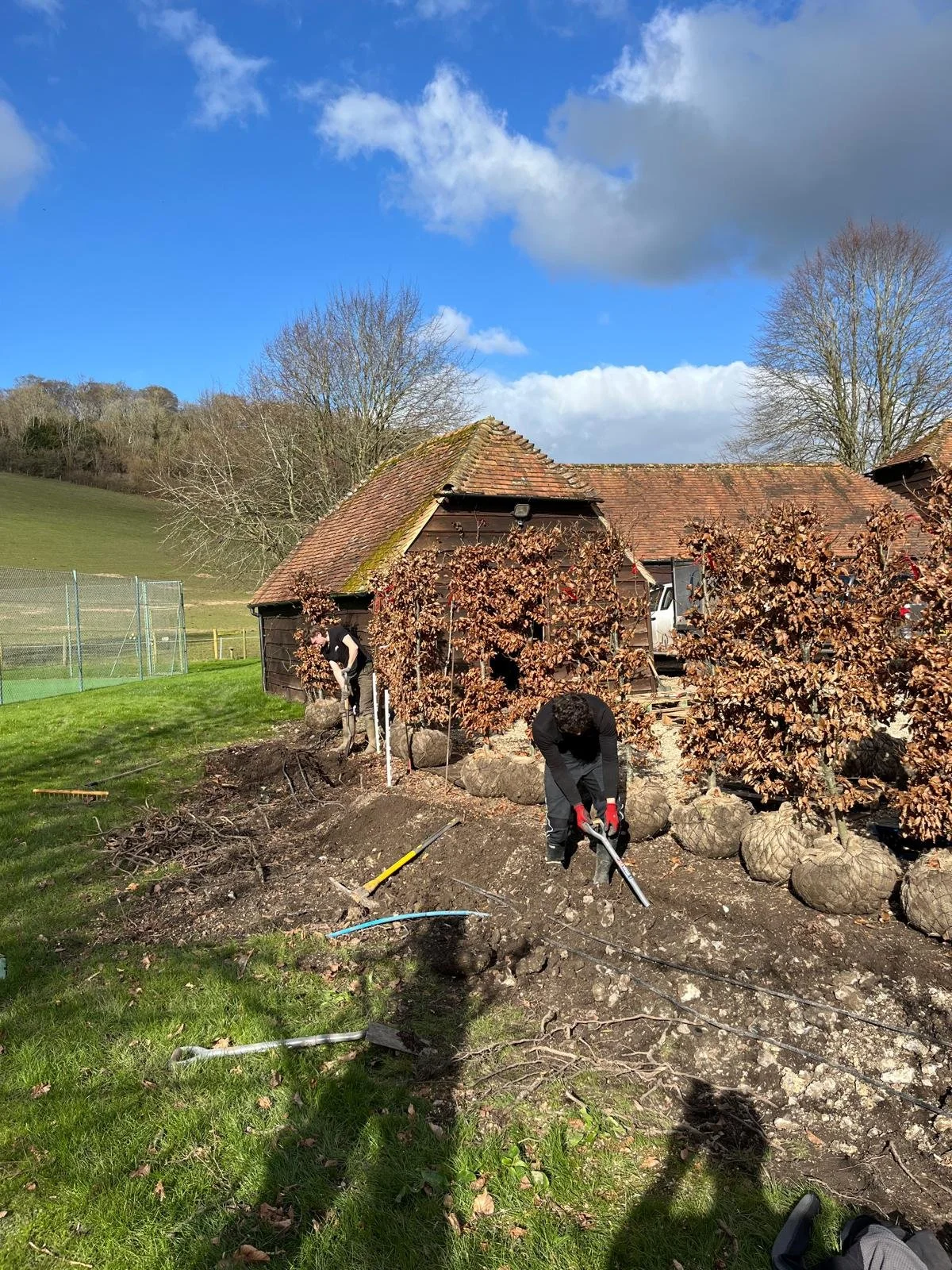 Two people working on digging and landscaping beside a small wooden house with a tiled roof. The scene is set outdoors on a bright, partly cloudy day with a grassy area and trees in the background.
