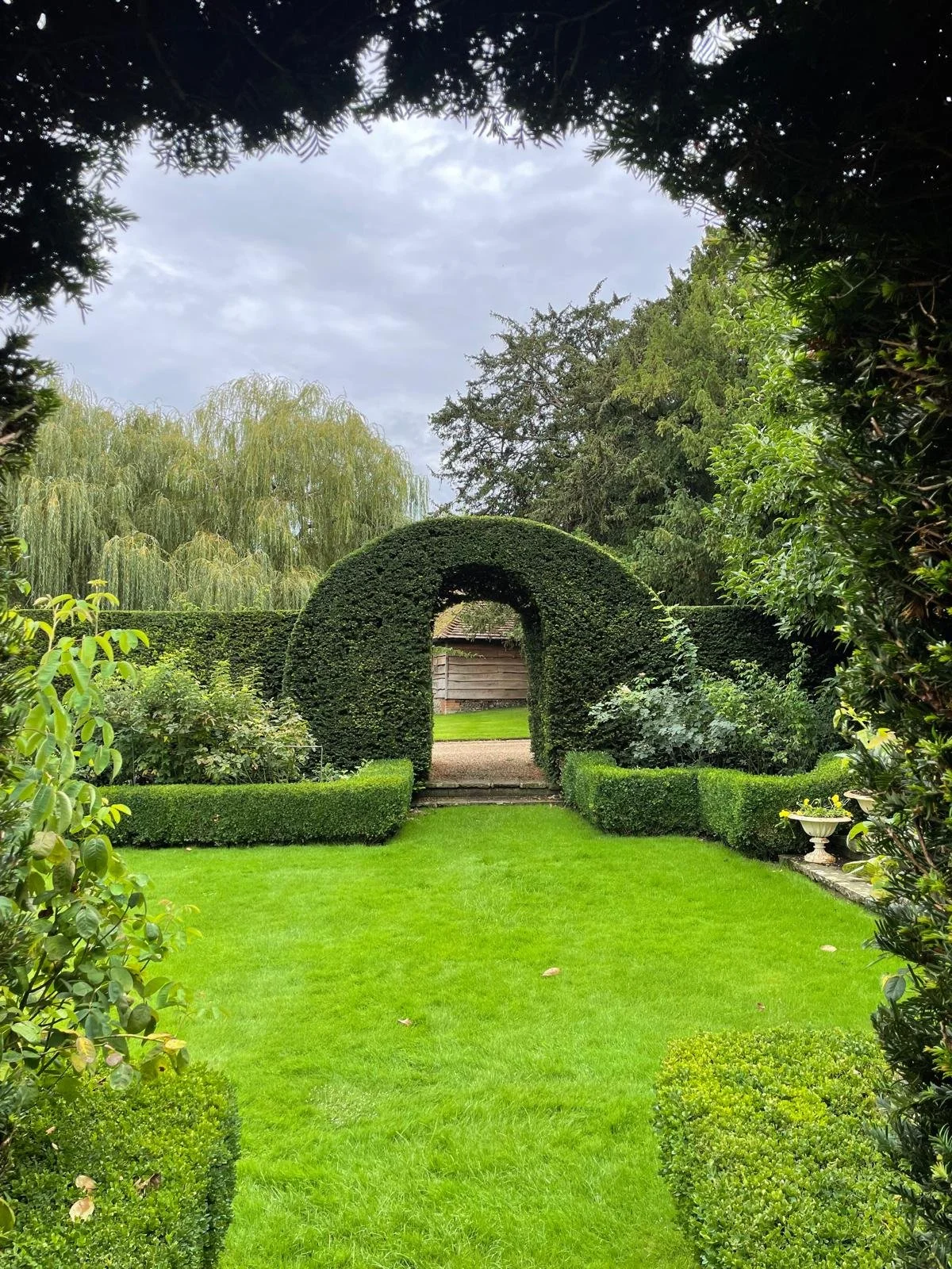 A well-maintained garden with lush green grass, trimmed hedges, and an arched topiary leading through to a gravel pathway and a wooden fence in the background, framed by trees and shrubs.