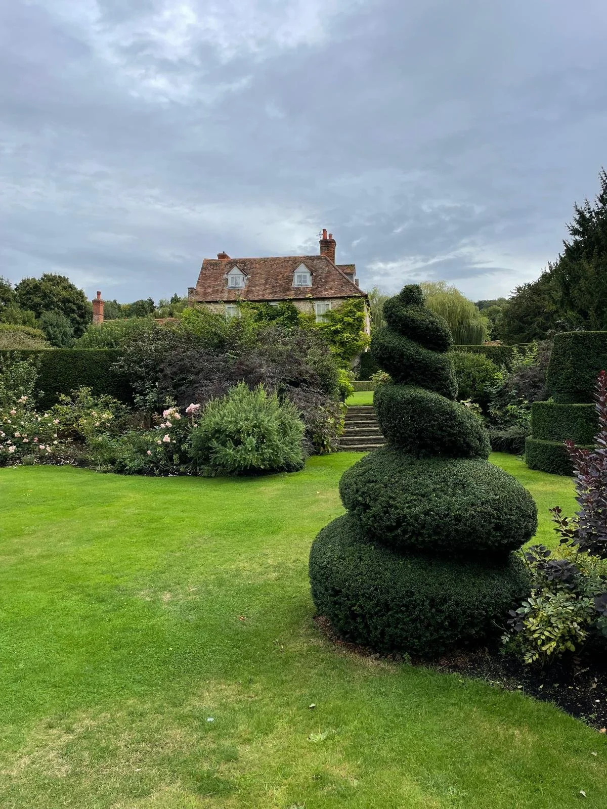 A landscaped garden with well-manicured grass, neatly trimmed bushes, and a house with a brick roof in the background under a partly cloudy sky.