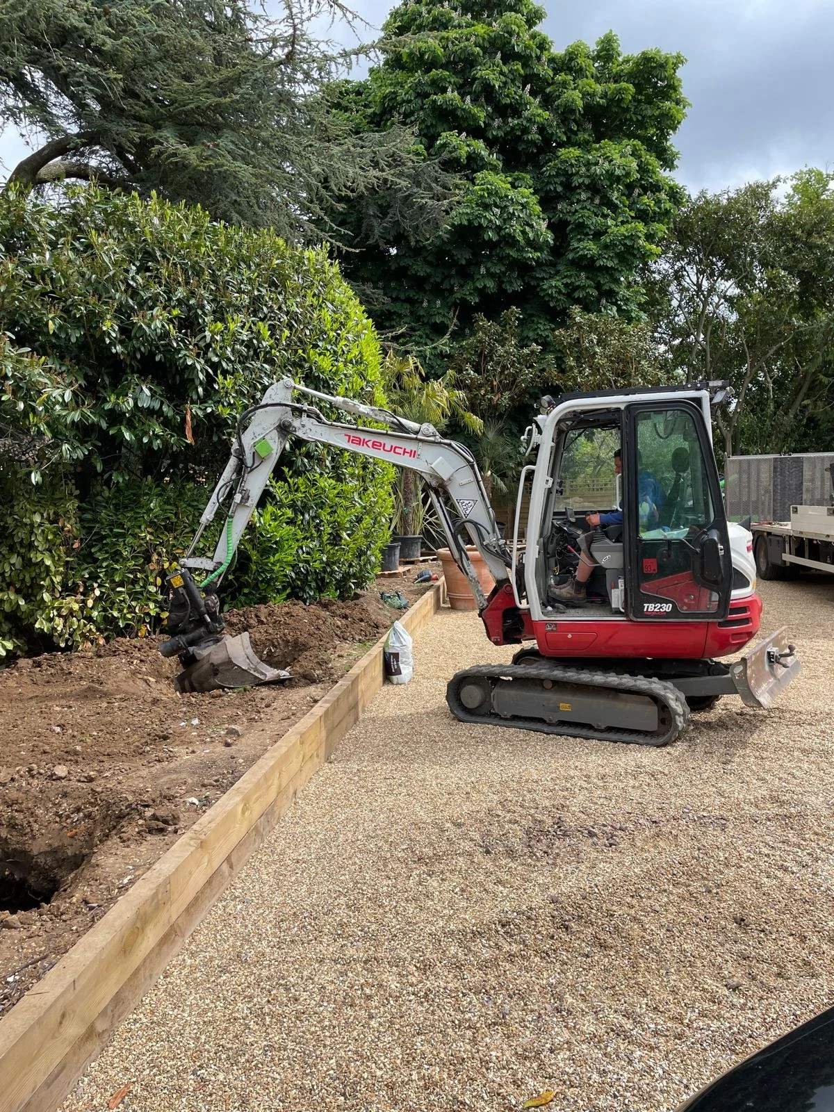 A small Takeuchi excavator digging a trench along a garden bed lined with a wooden barrier. The excavator is operated by a person seated inside the cab, wearing a blue shirt. There are lush green trees and bushes in the background, along with a cloud