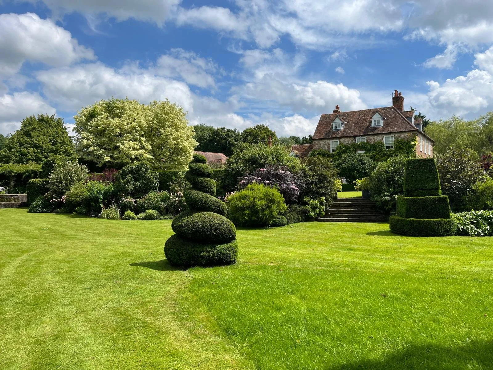 A lush green garden with manicured lawn, topiary bushes, and a large house in the background under a partly cloudy sky.
