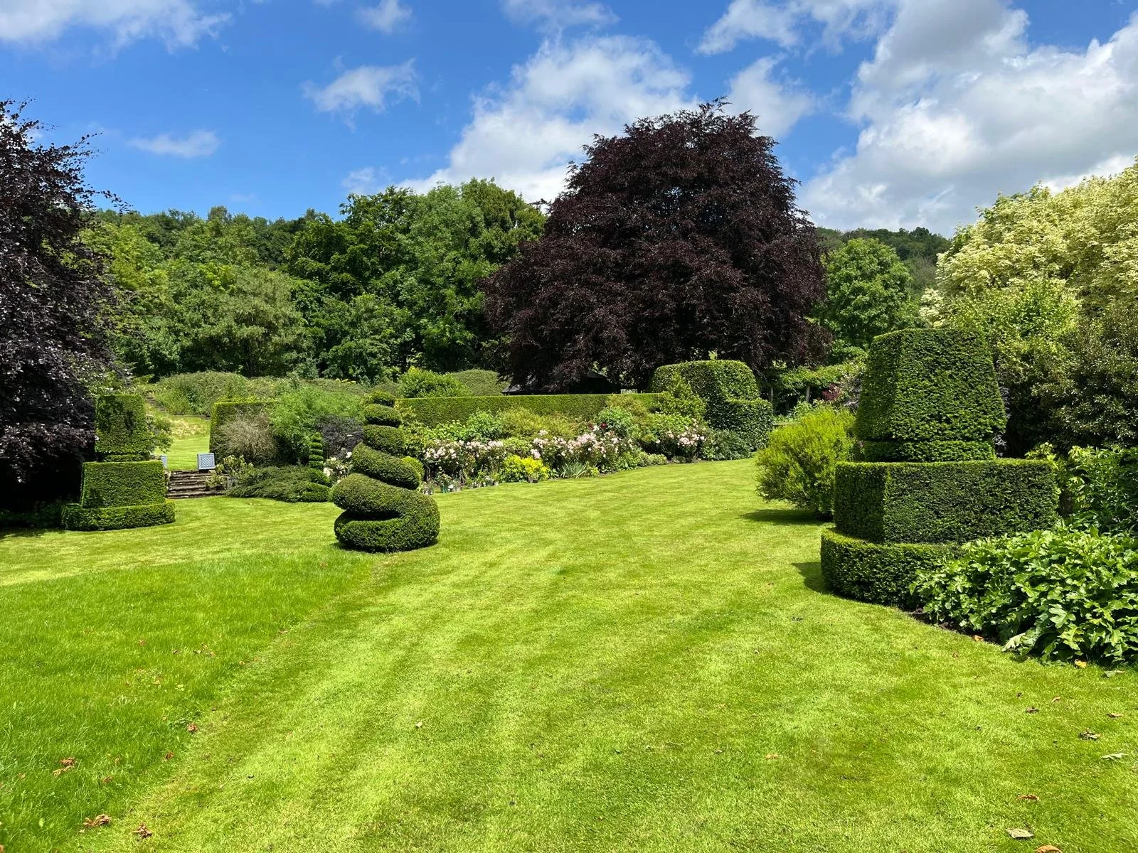 A well-manicured garden with trimmed bushes and trees, a green lawn, blooming flowers, and a clear blue sky with some clouds.