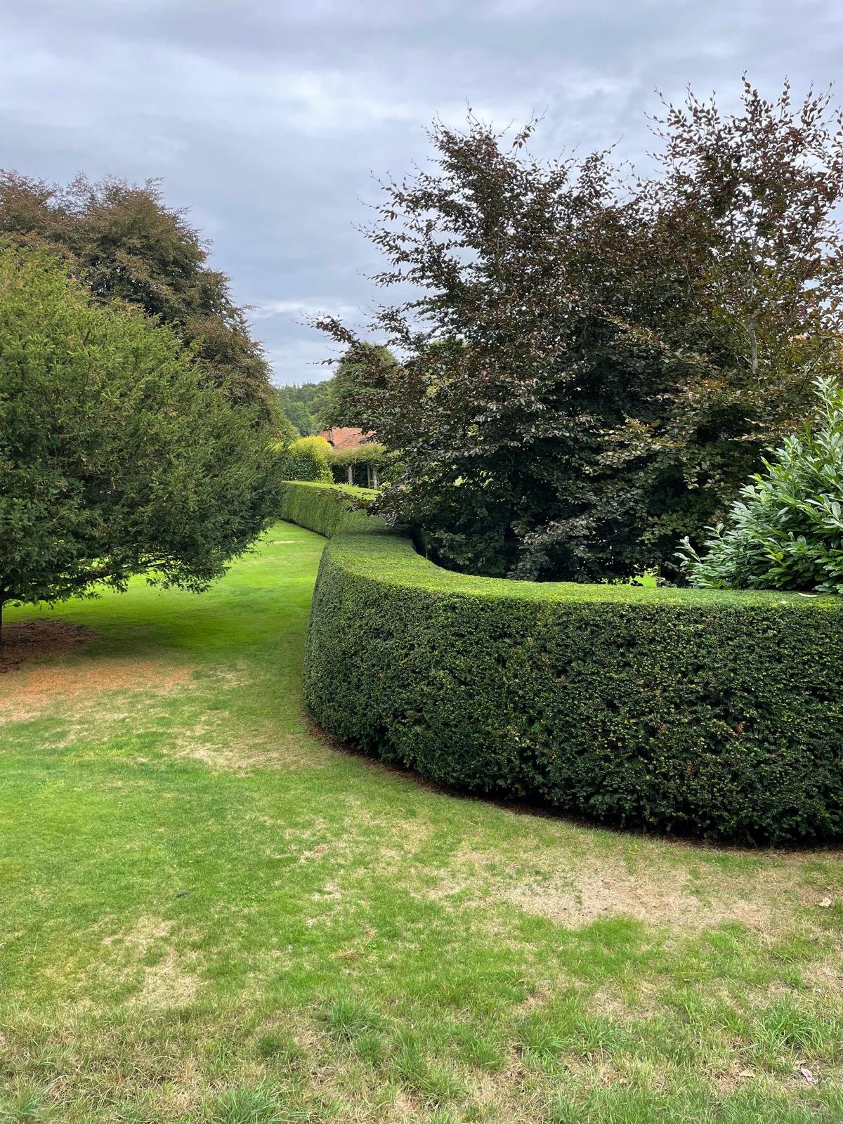 A well-maintained park with green grass, trimmed hedges, and trees, under a cloudy sky.