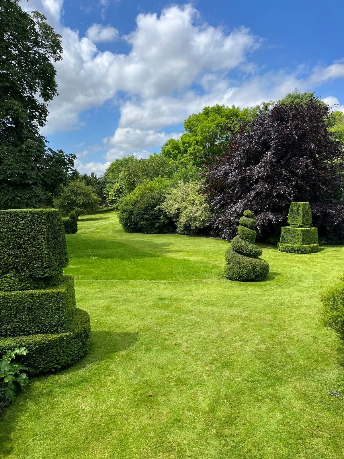 Well-manicured garden with trimmed bushes, topiary, and lush green grass under a blue sky with clouds.