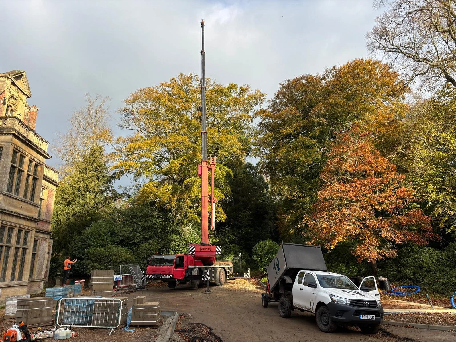 Construction site with trees showing fall colors, a large crane, a white utility truck, and a person in a safety vest operating equipment.