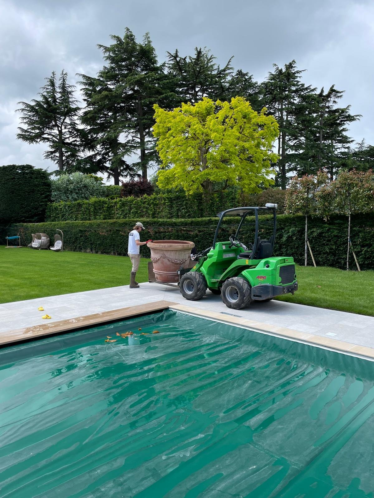 A person working with a green mini loader near a swimming pool and large plant pot outdoors on a patio, with trees and cloudy sky in the background.