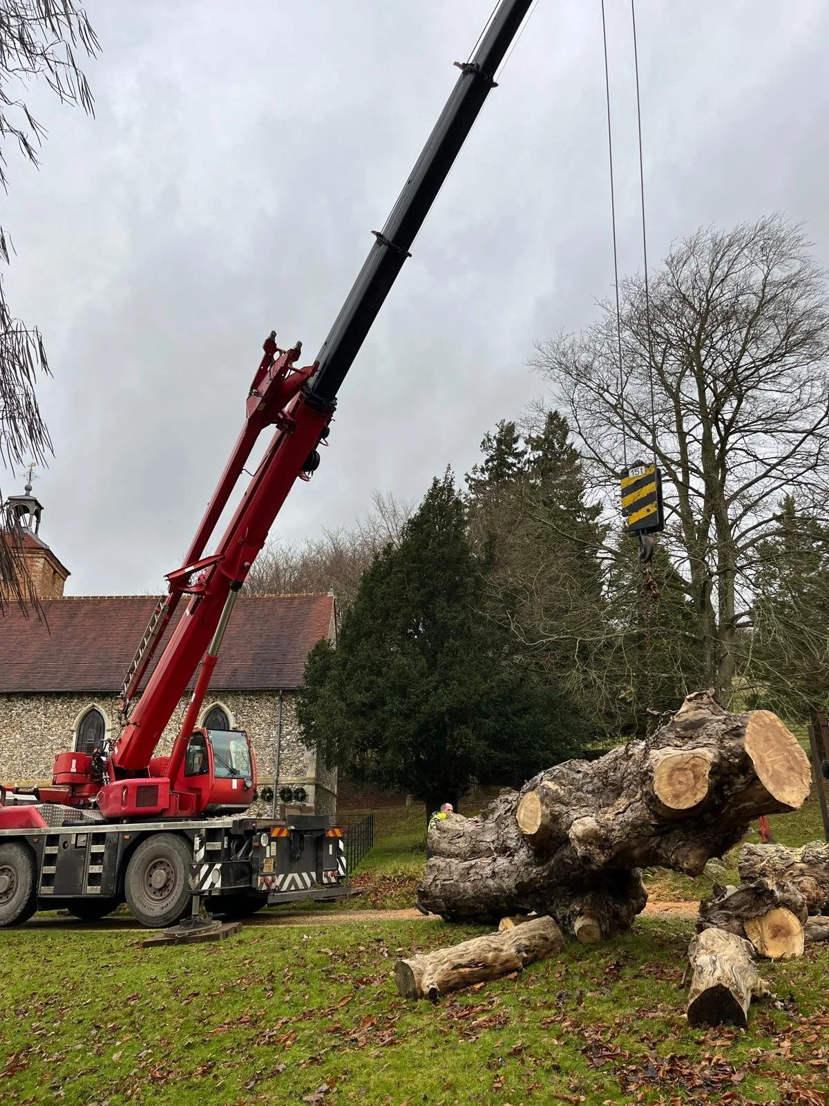 A red crane lifting a large tree trunk log in a grassy area with trees and a stone church building in the background, overcast sky.