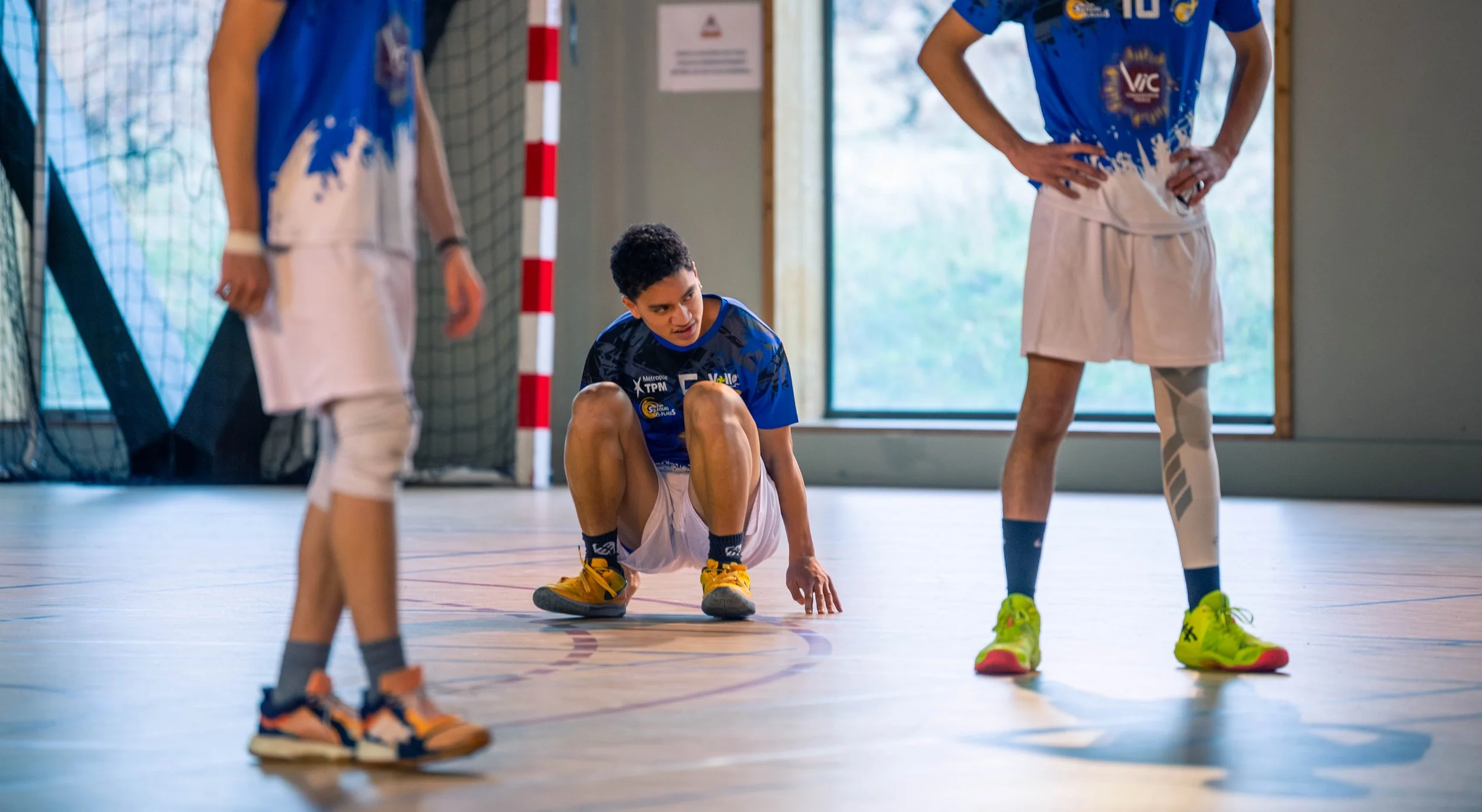 Jeune garçon en position accroupie sur un terrain de sport intérieur, entouré de deux autres garçons. Tous portent des vêtements de sport bleus et des chaussures de sport colorées.