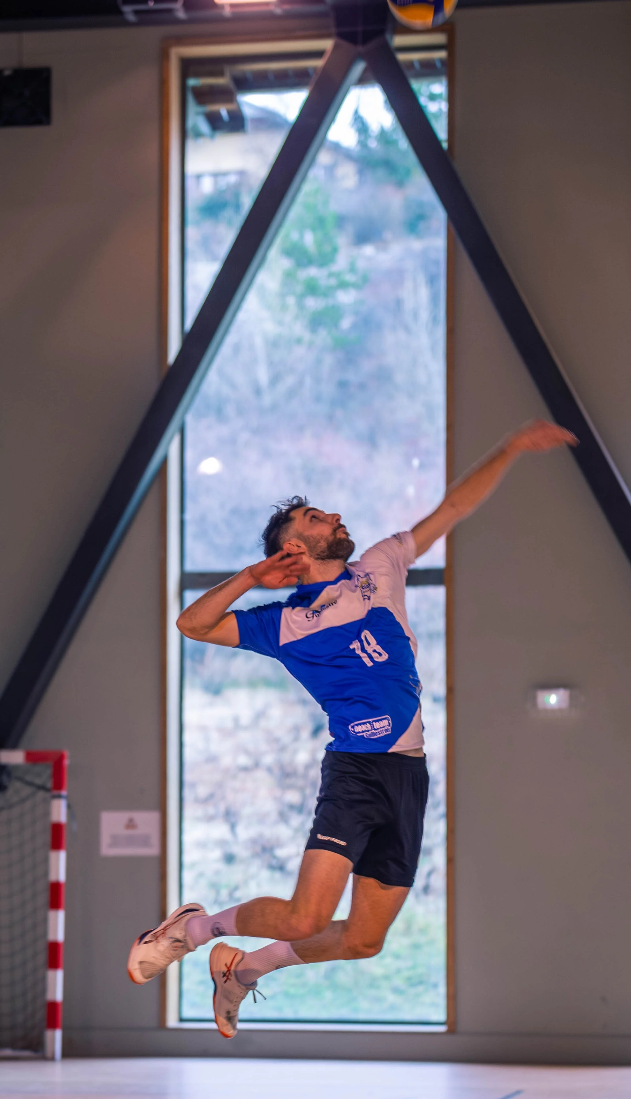 Un homme en maillot de sport bleu et noir saute dans une salle d'intérieur, avec un grand fenêtre derrière lui.