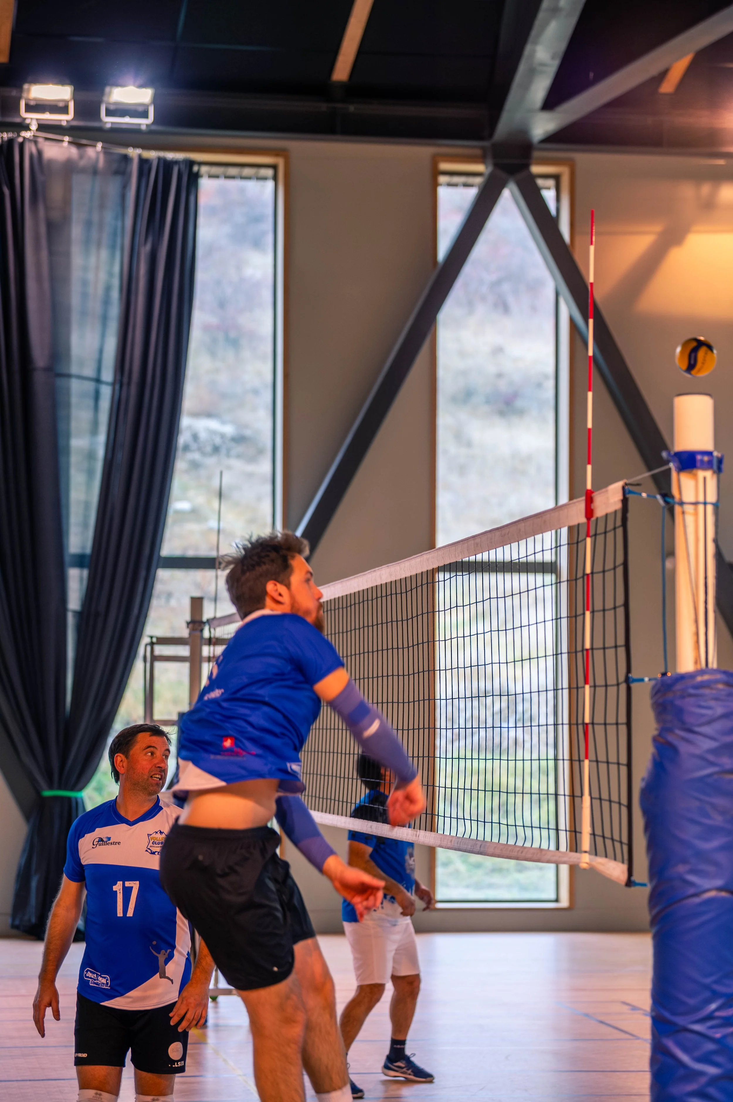 Des hommes jouent au volleyball intérieur, un joueur en train de sauter pour frapper le ballon vers le côté adverse, avec un filet au centre.