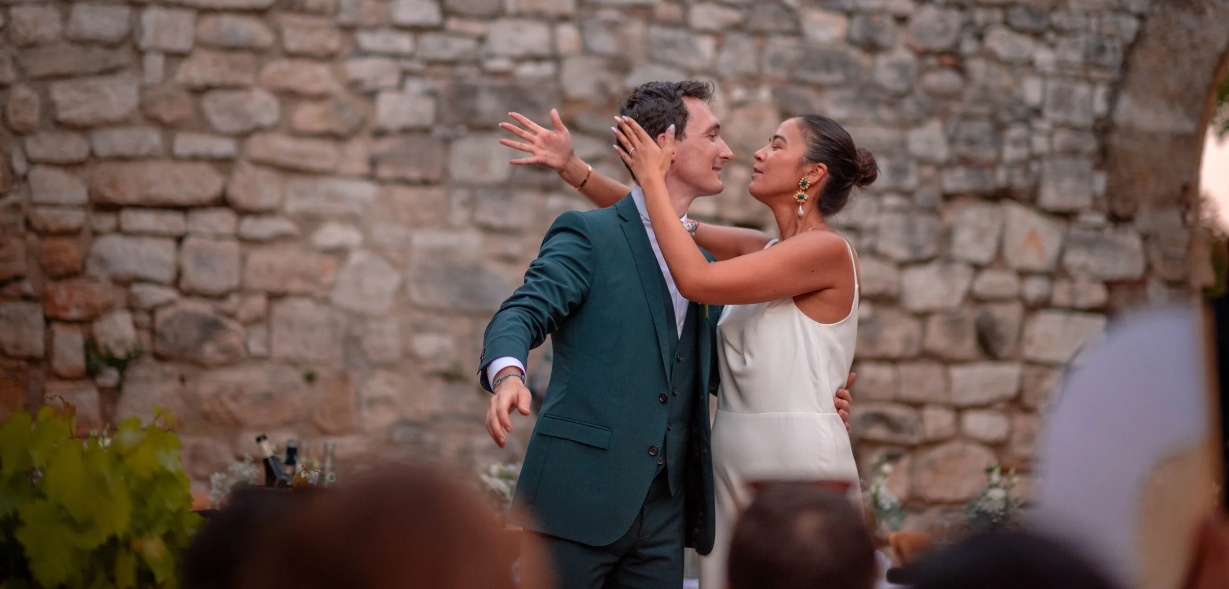Un couple danse lors d'un événement en extérieur devant un mur en pierre, la femme porte une robe blanche et des bijoux, l'homme porte un costume vert foncé.