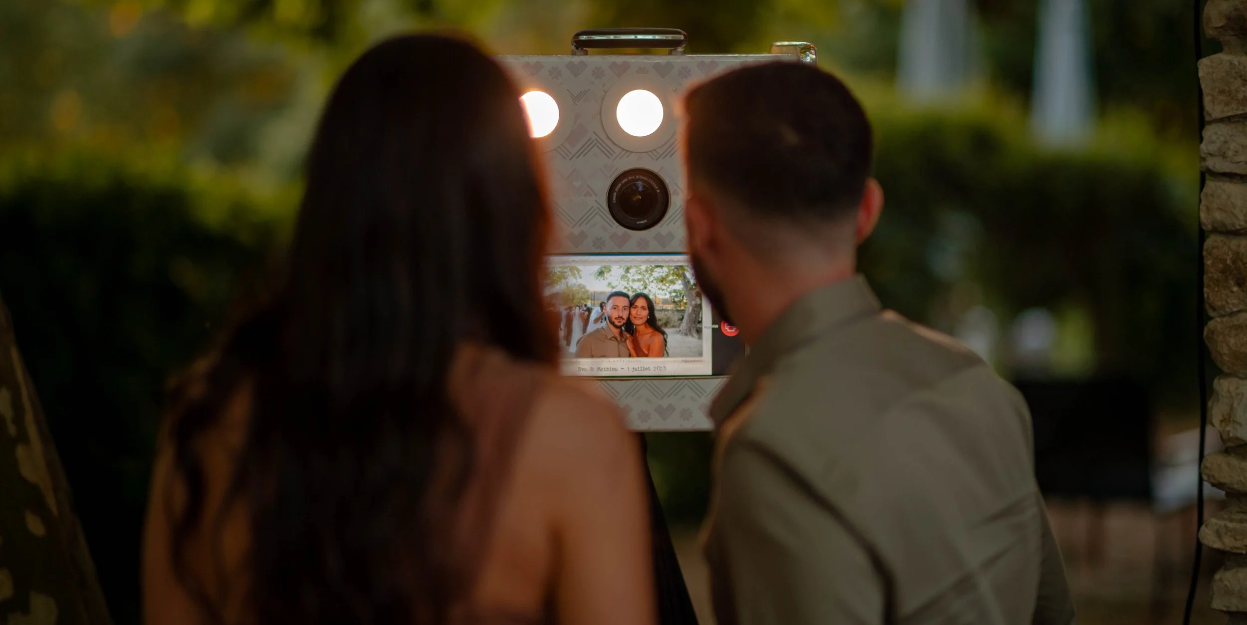 Un couple regarde une photo sur un photobooth lors d'un événement en soirée, avec un décor extérieur entouré de verdure.